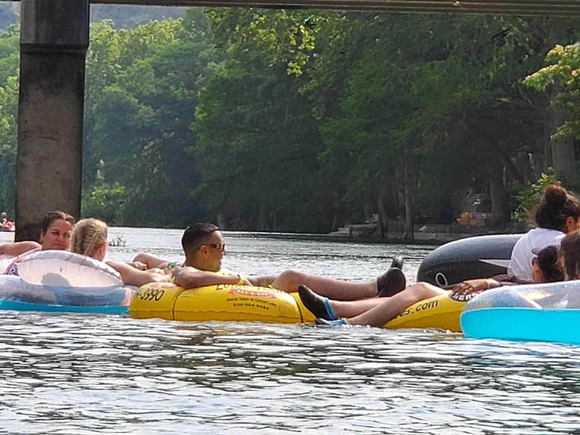 People floating on inner tubes in a river. A man in a yellow tube rests with a drink; trees line the banks under a bridge.