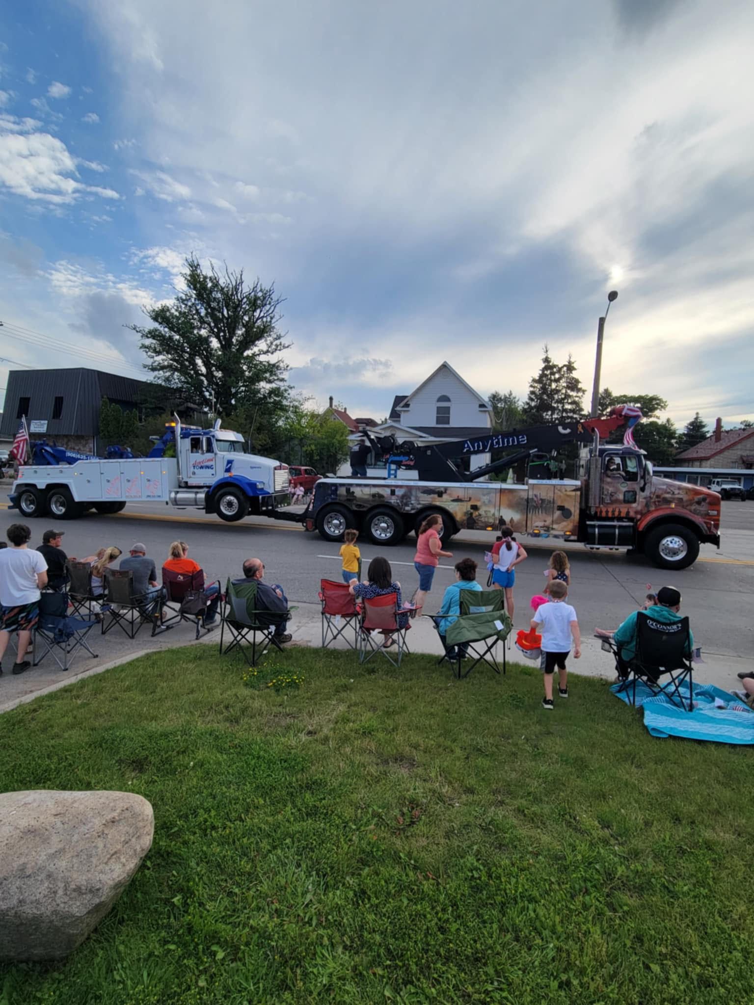 Tow trucks in a parade, people watching from lawn chairs on a grassy area under a cloudy sky.