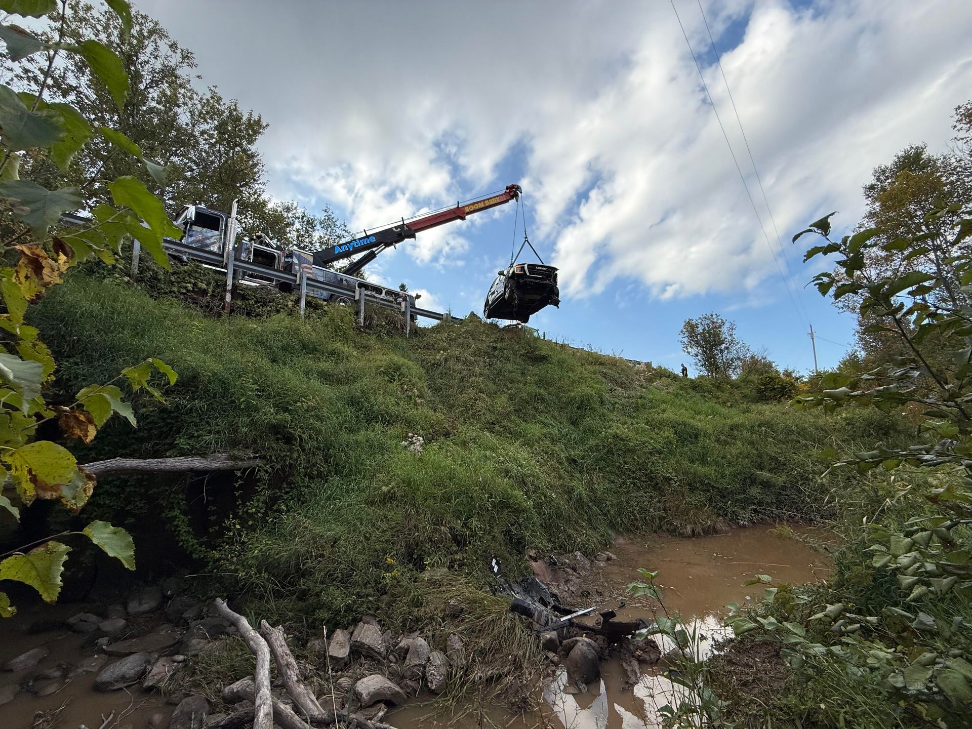 A crane lifting a dark object over a muddy bank. The scene is outdoors with green foliage and a partly cloudy sky.