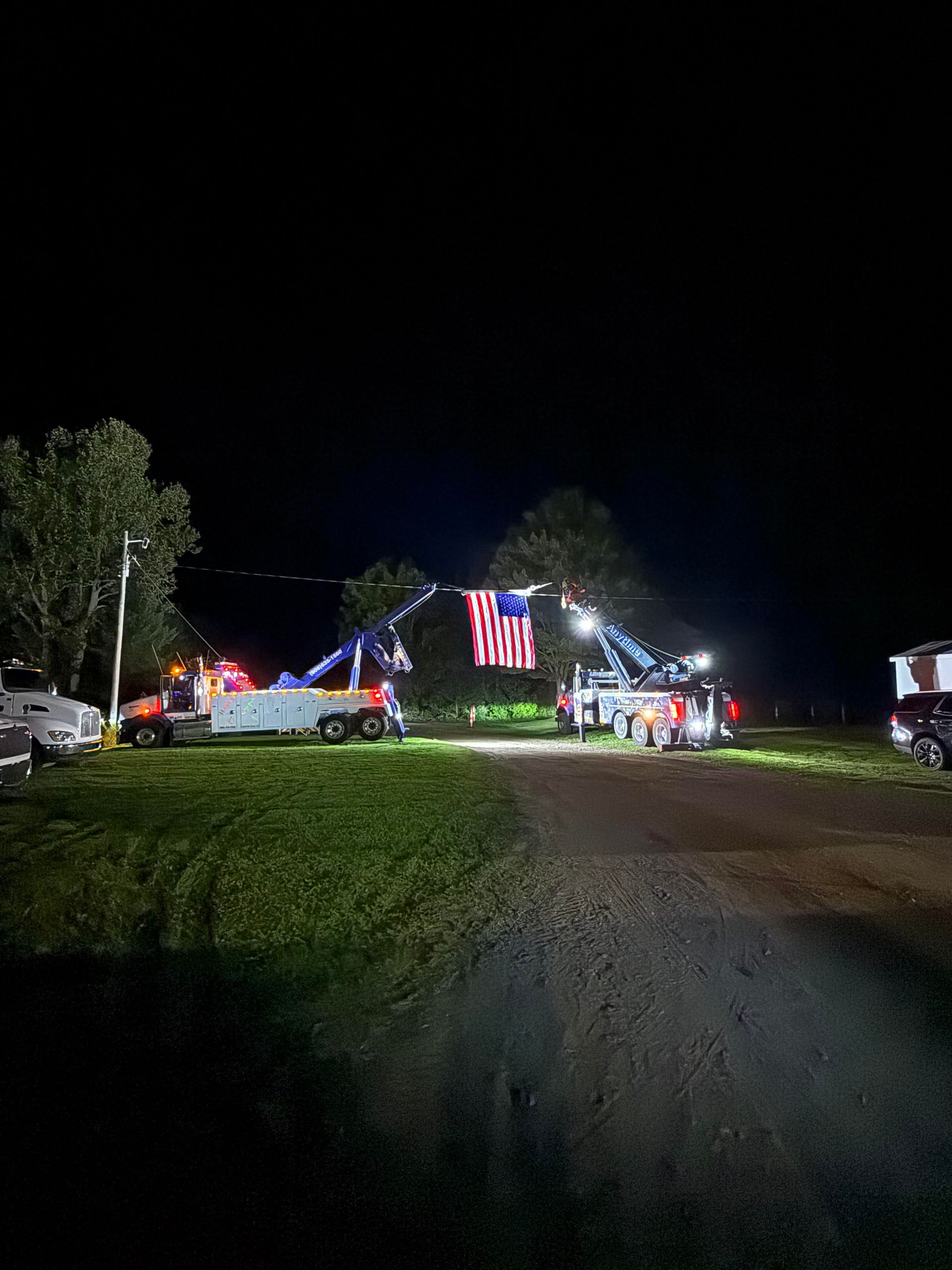 Two tow trucks with American flag suspended at night on grassy area.