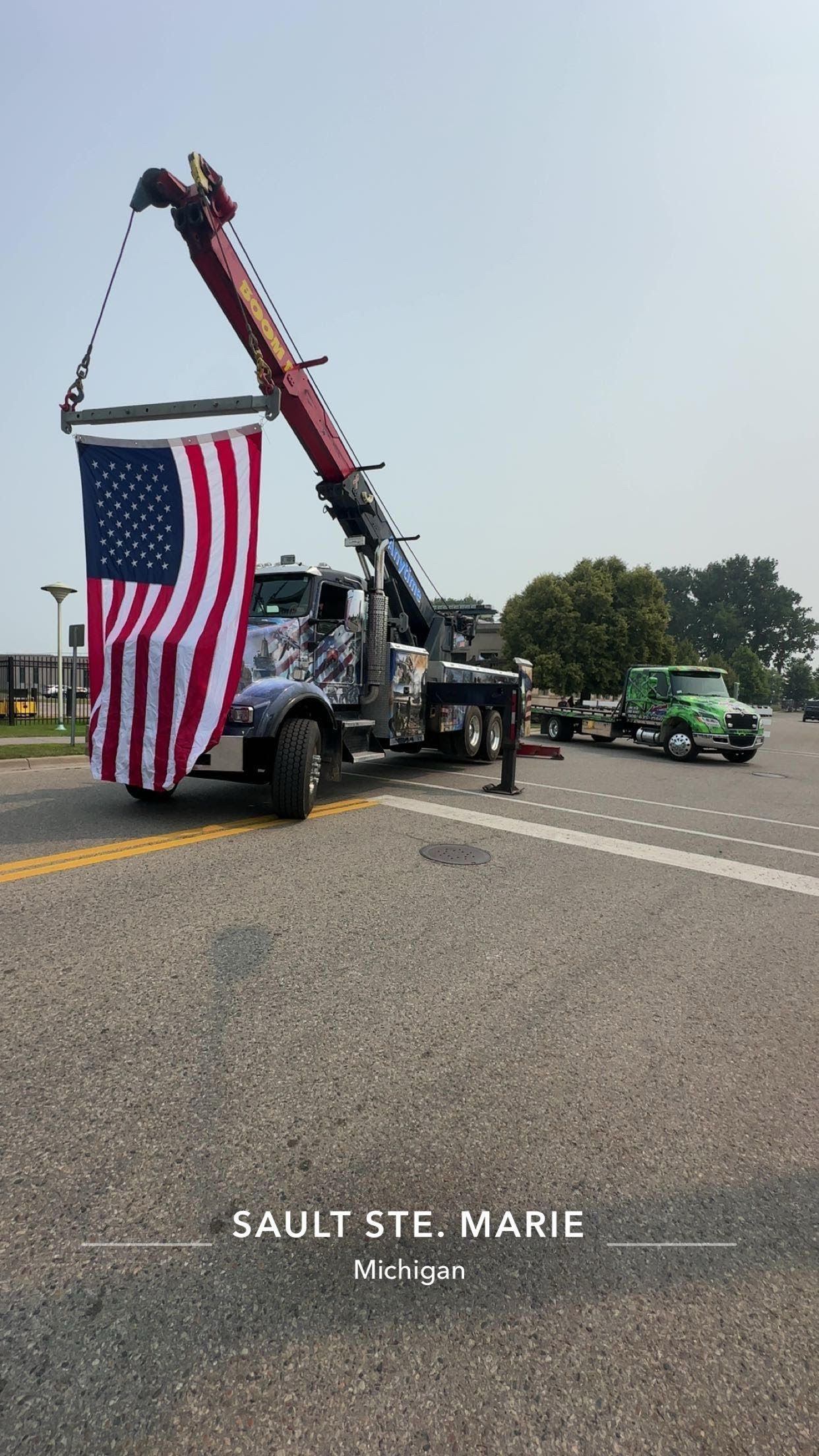 Tow truck with extended crane holding an American flag; a green truck is parked nearby.
