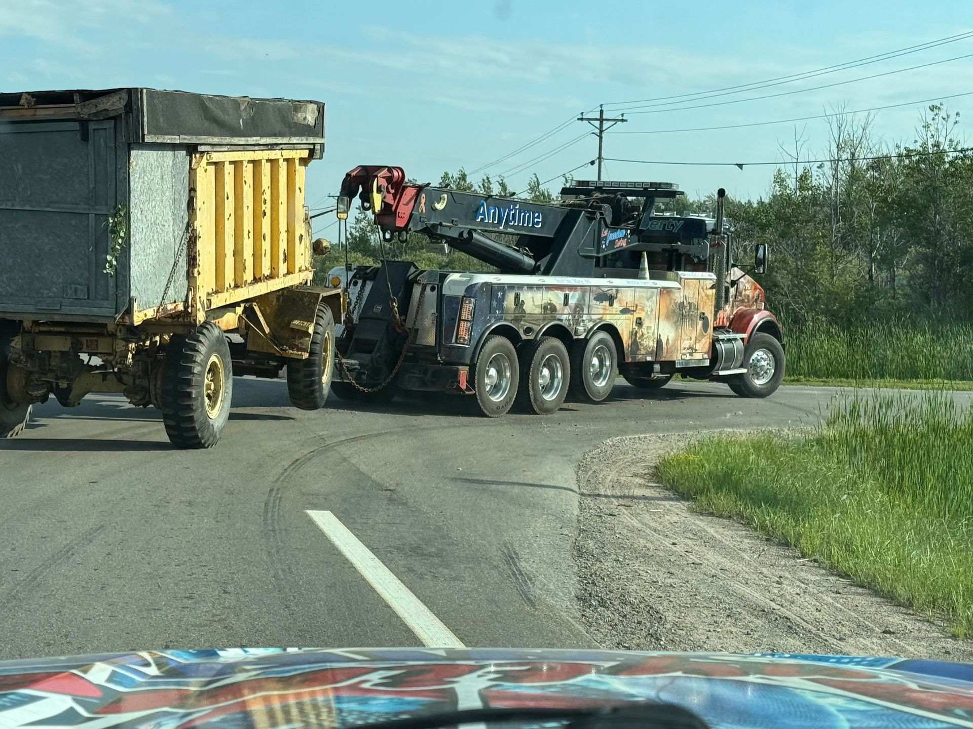 Tow truck towing a yellow dump truck on a road with green grass and a blue sky.