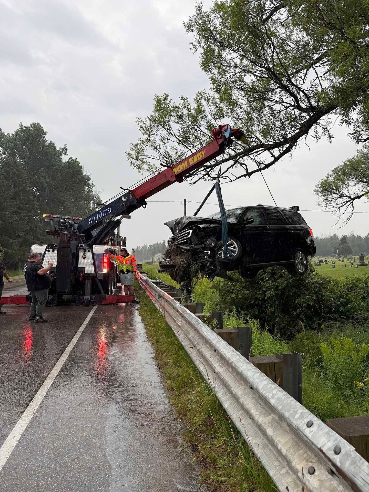 Tow truck lifting a badly damaged black vehicle from a ditch beside a road