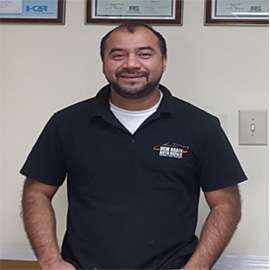 Man wearing black polo shirt with logo, standing in front of light-colored wall, smiling.