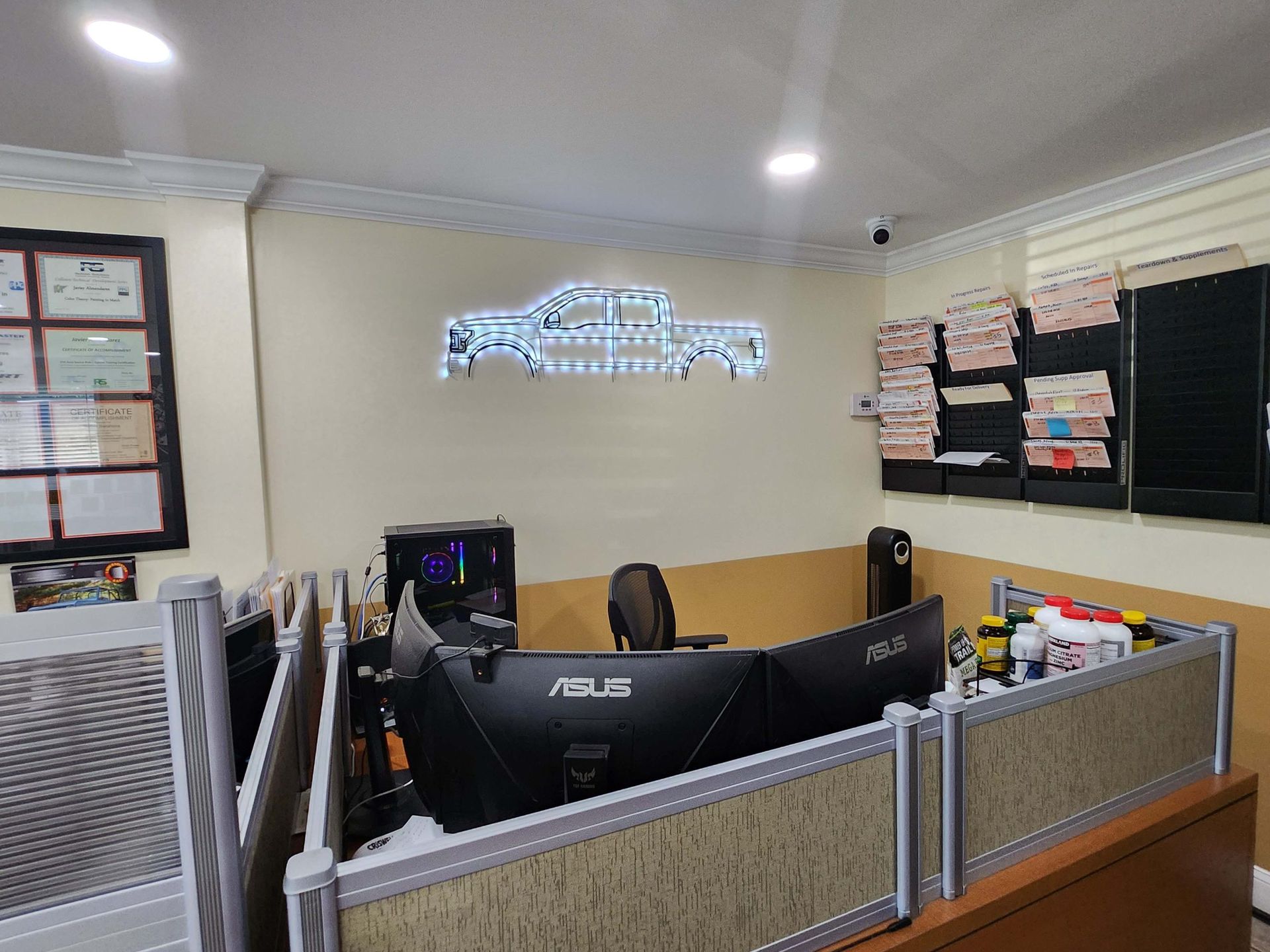 Office with computer screens, decorative truck outline sign, cubicle dividers, and various items on desks.