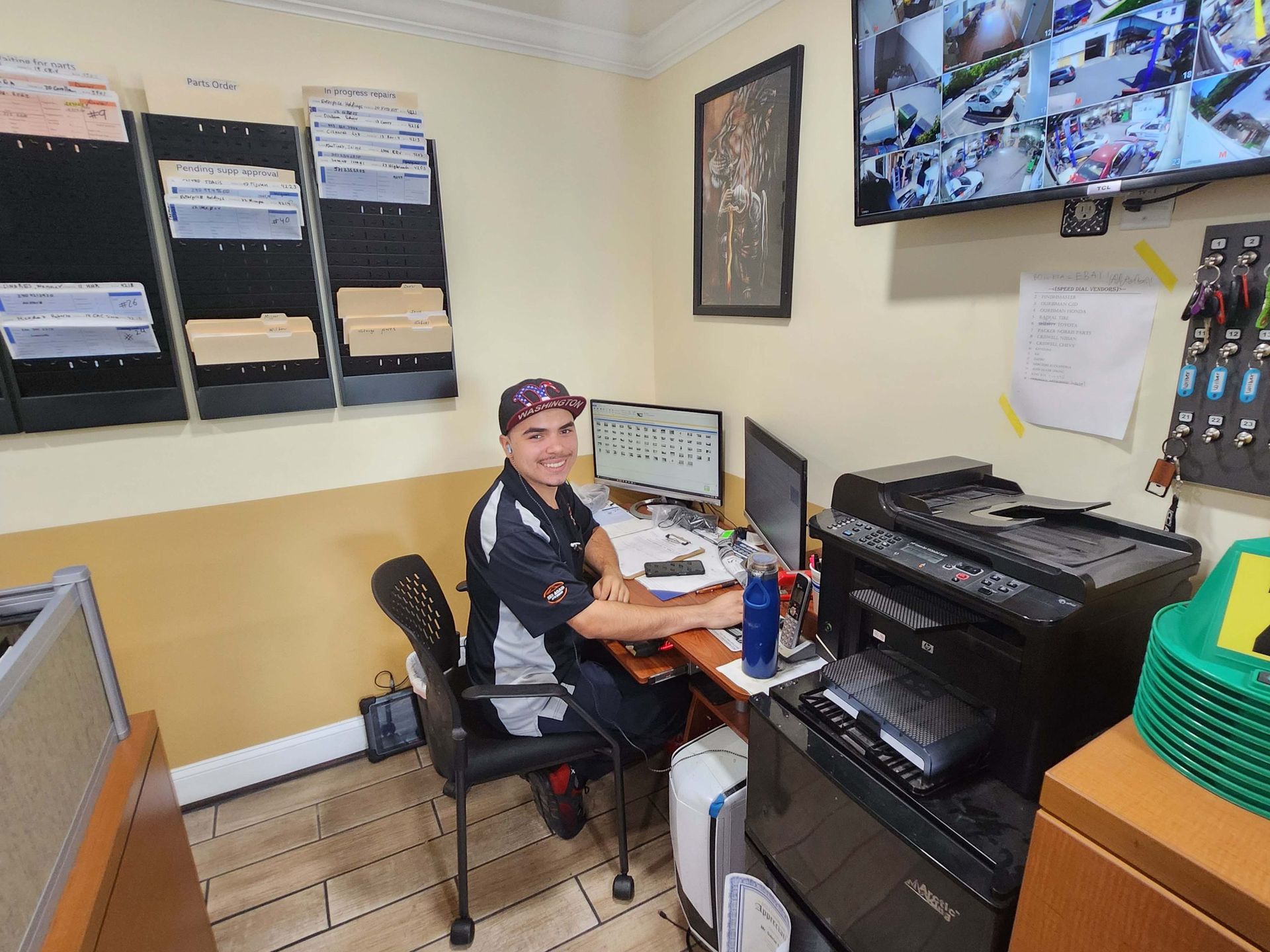 Person in a shop uniform at a desk, smiling. Monitors, printer, and paperwork visible. Security cameras and a wall of keys are in the background.