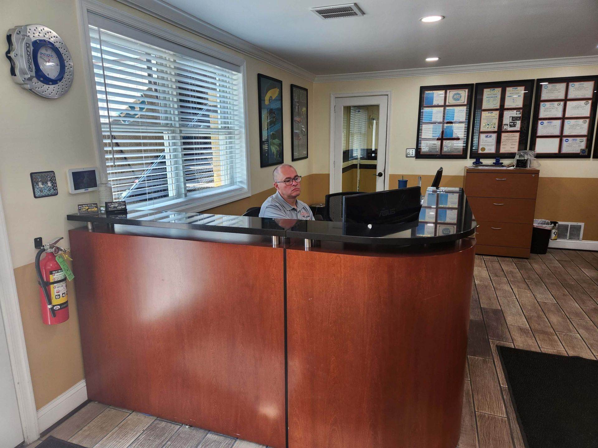 Reception area with a person at the desk, looking at a computer monitor. Brown wood desk, certificates on wall.