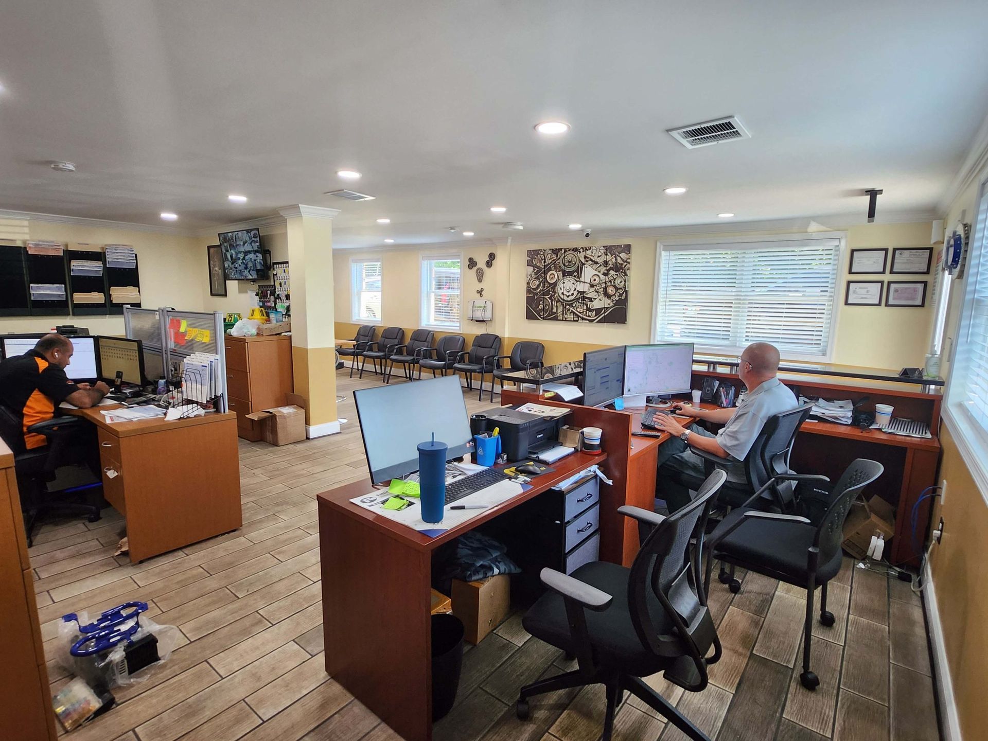 Office with several desks, computers, and people working. Chairs and waiting area visible.