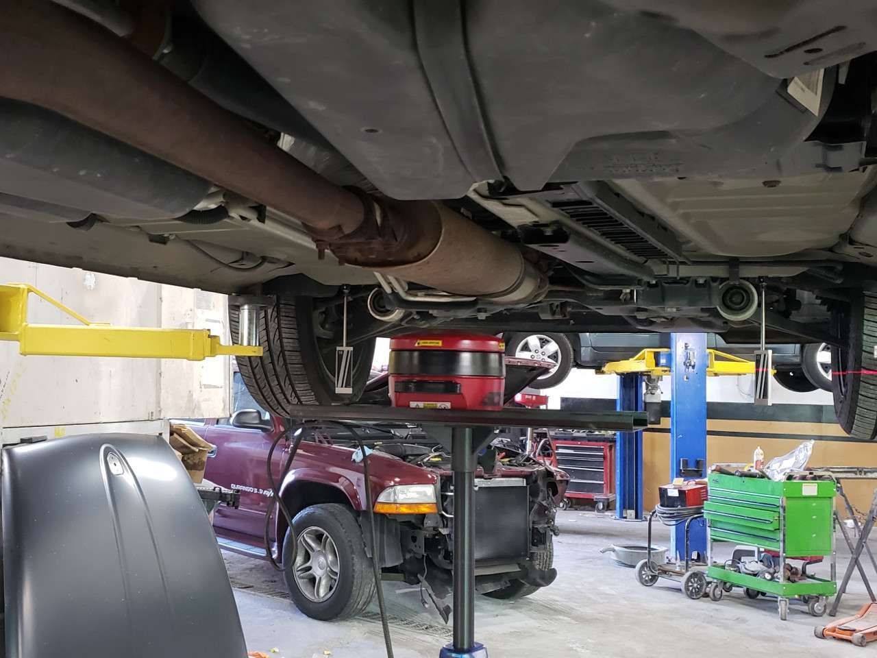 Car on a lift, exposing exhaust and undercarriage, in a repair shop. Another car is being worked on below.