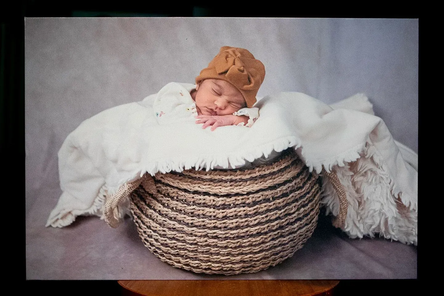 Newborn baby wearing a brown hat, nestled in a woven basket with a white blanket, resting on a gray background.