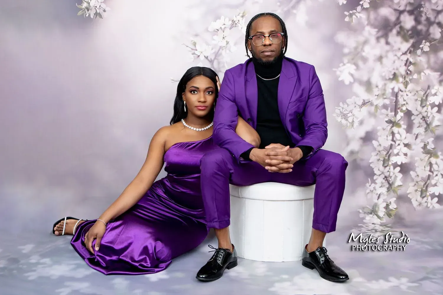 Couple poses; woman in purple dress, man in purple suit seated on white cylinder, floral backdrop.