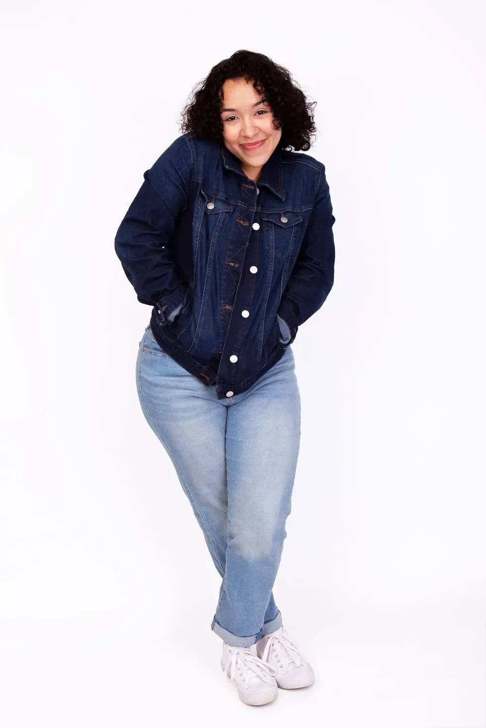 Woman in a denim jacket and jeans, hands in pockets, smiling. White background.