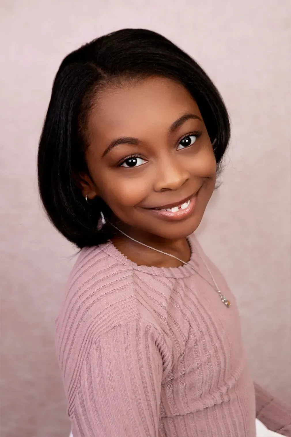 Girl with short, dark hair smiles, wearing a pink textured long-sleeved shirt, soft background.