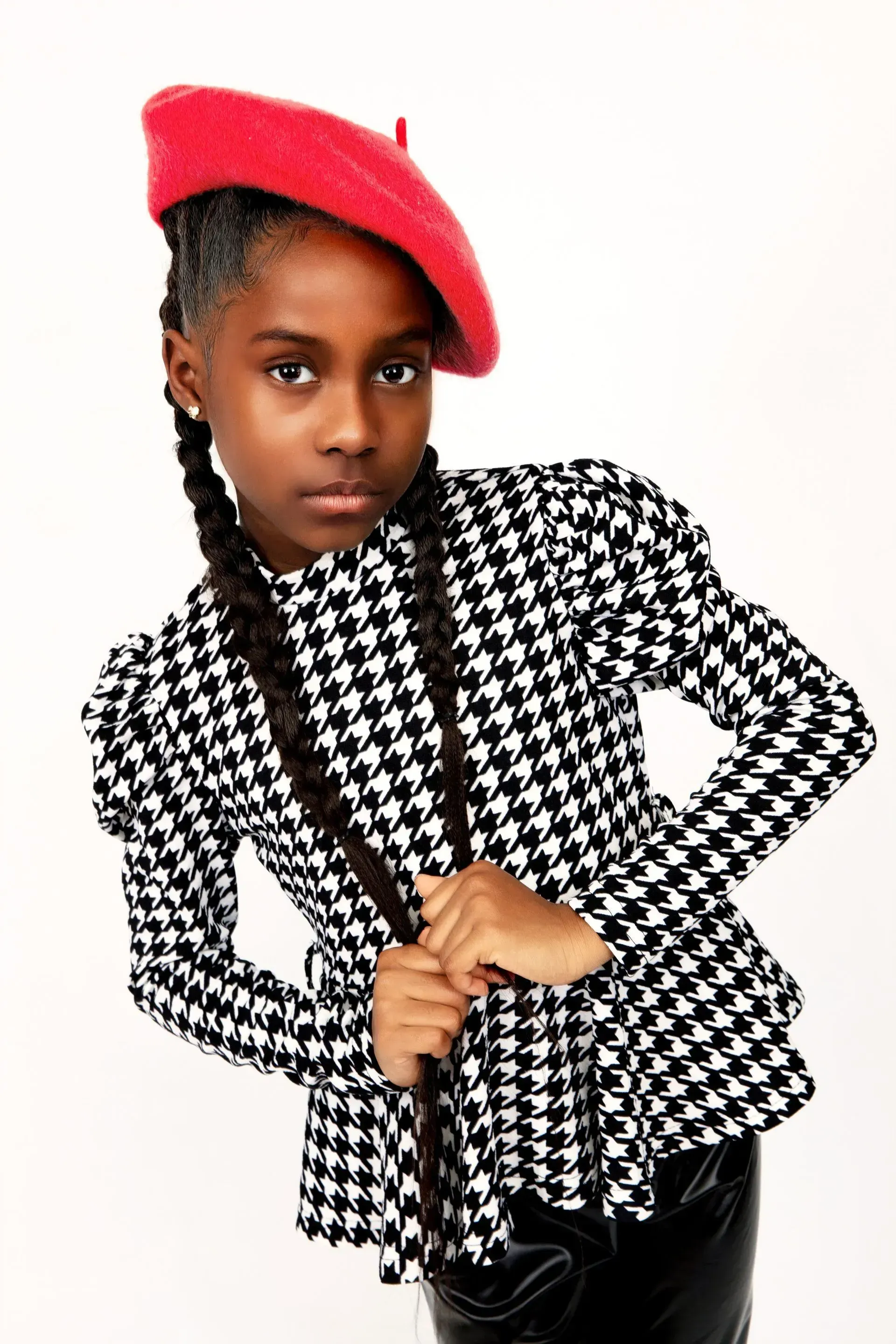 Girl wearing red beret and houndstooth jacket, holding braids, against a white background.