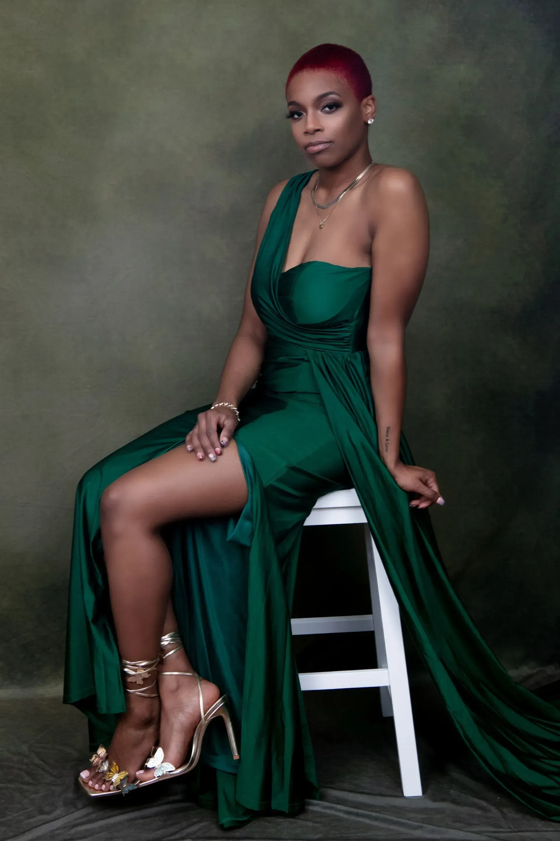 Woman in a green formal dress sits on a white stool, looking at the camera. She has short red hair and wears gold heels.