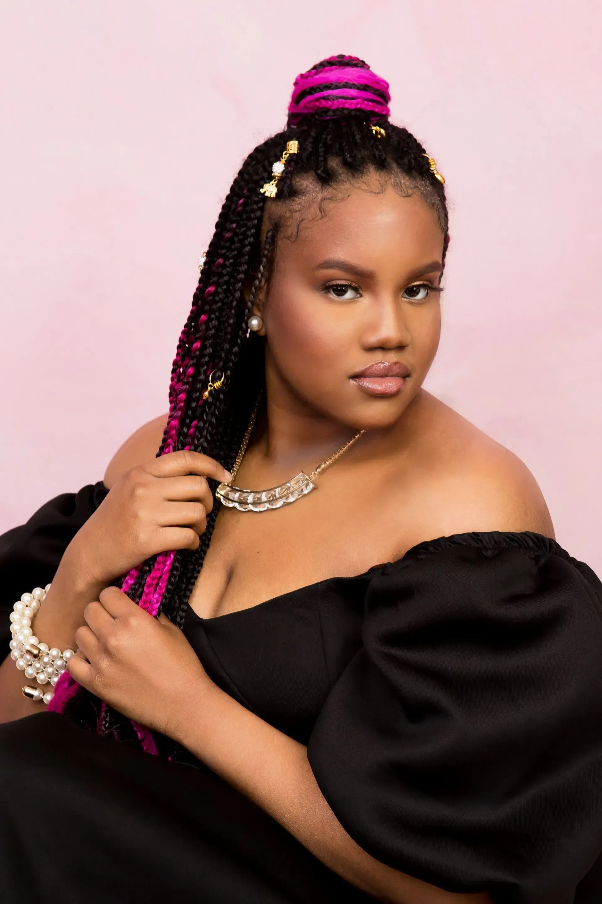 Woman with braids, pink highlights, in an off-the-shoulder black dress, looking at the camera.
