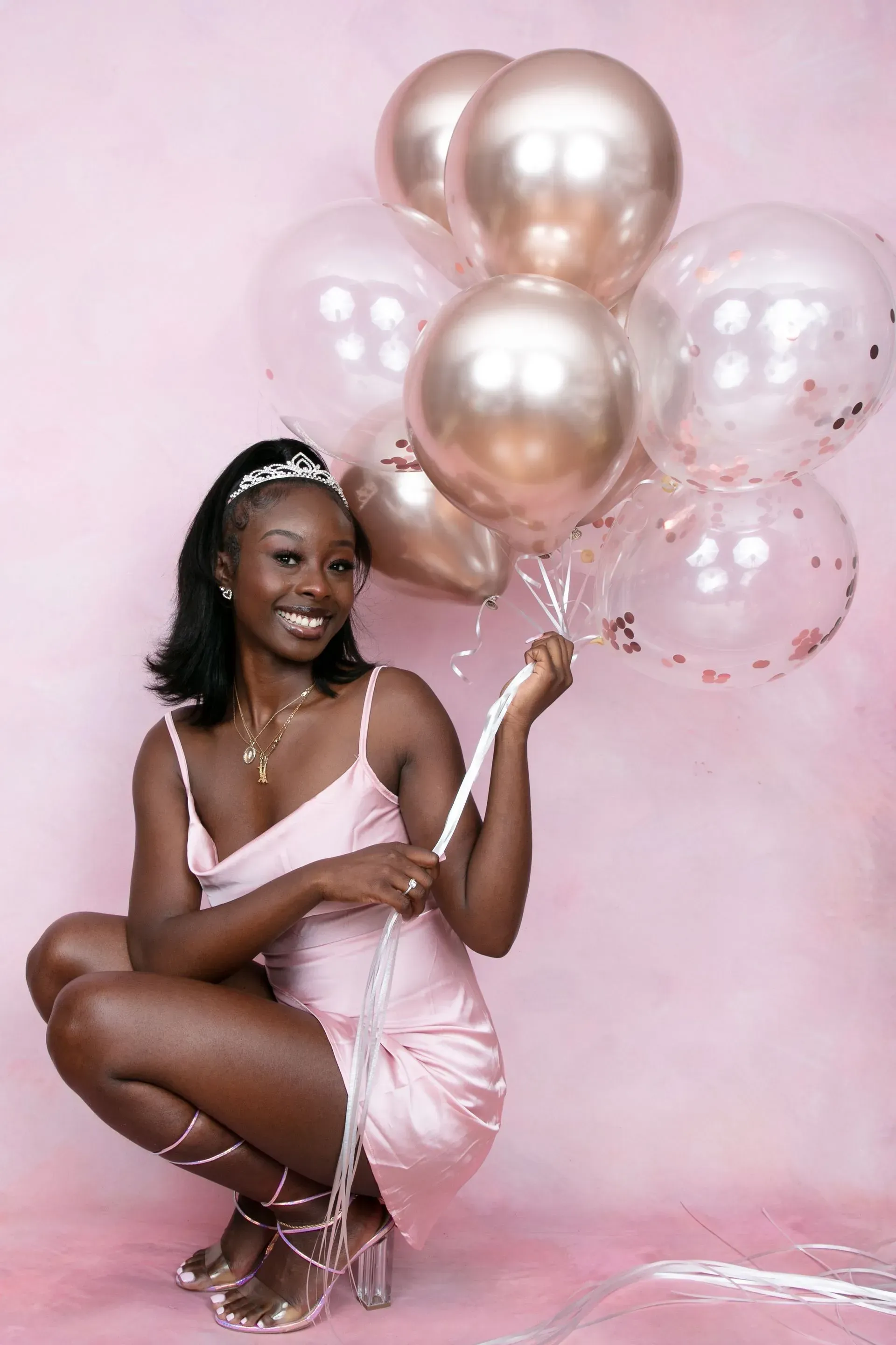 Woman holding balloons, wearing a pink dress and tiara, smiling, squatting against a pink backdrop.