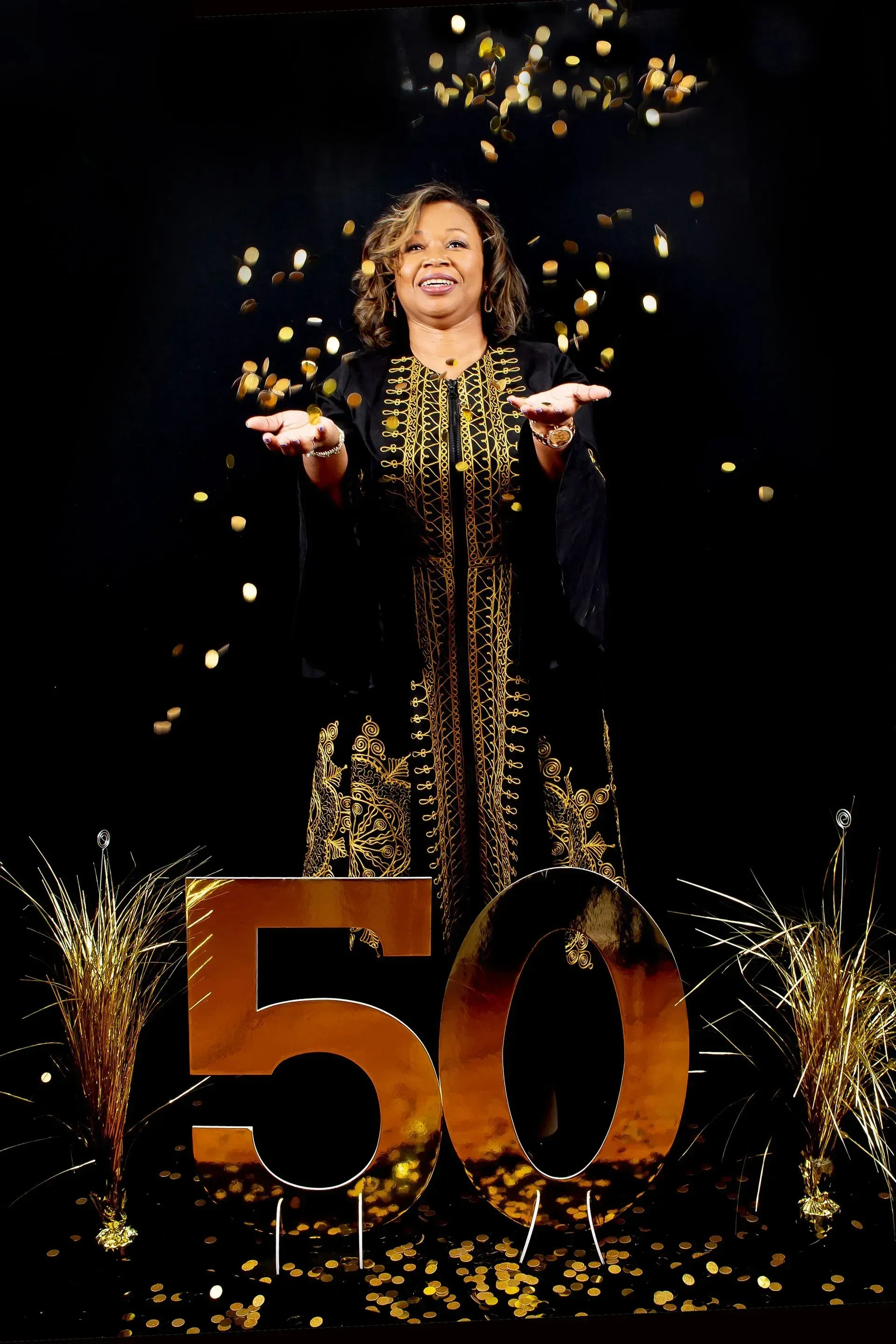 Woman in gold-trimmed black robe celebrating 50th birthday, throwing confetti. Black background with gold decorations.