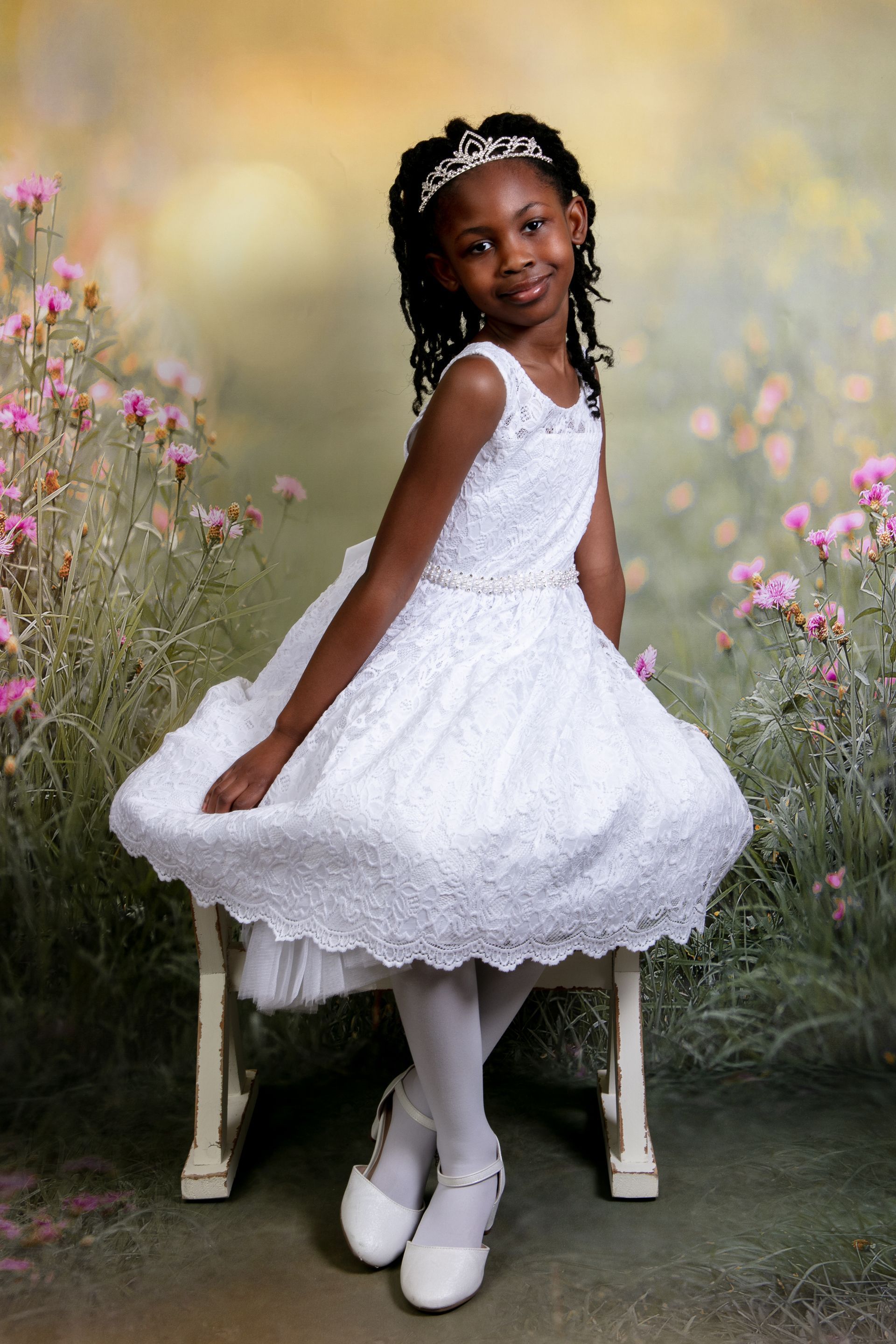 A person in a white lace dress and tiara sits on a chair in a field of flowers with a soft, painted-style background.