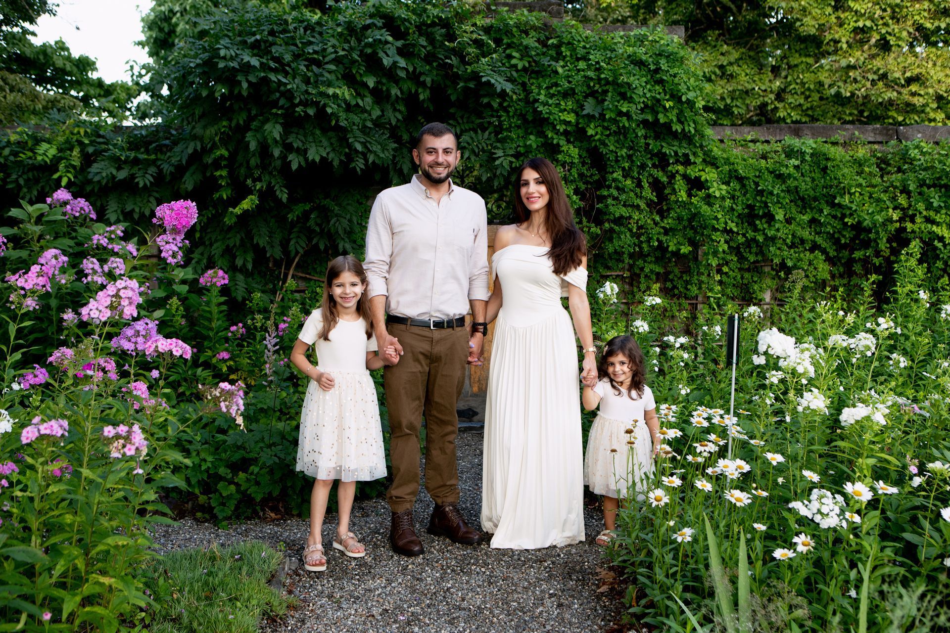 A family of four stands together on a gravel path in a lush, blooming garden.