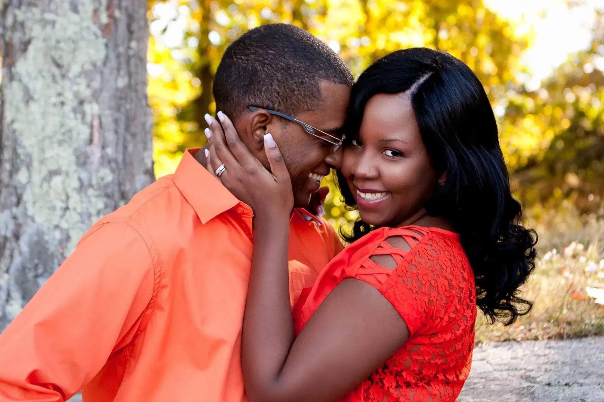 Couple embraces outdoors, smiling. Woman in red dress touches man's face, both wearing orange shirts.
