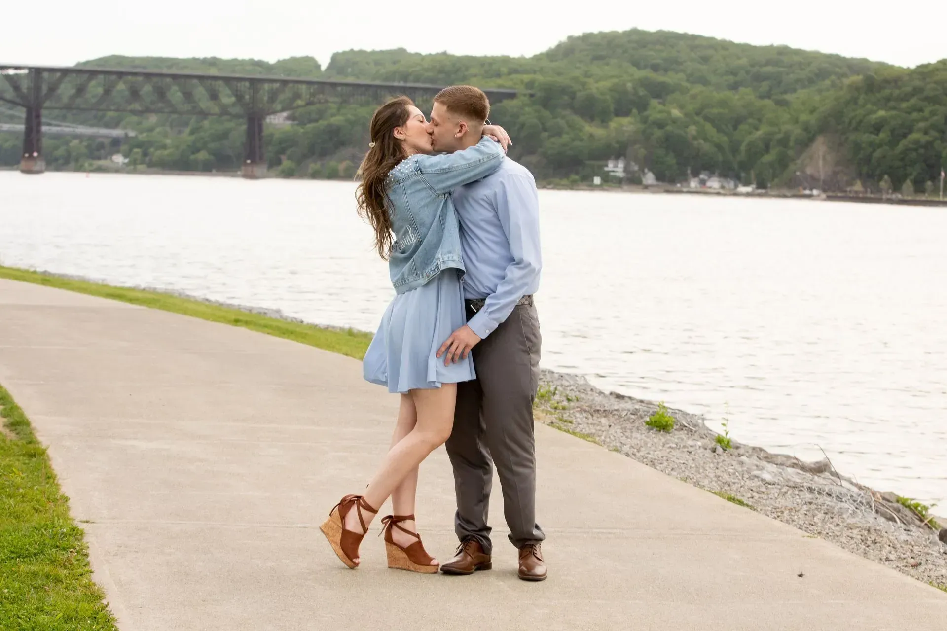Couple kissing on a path by a river, bridge in background. She wears a blue dress, he wears a light blue shirt.