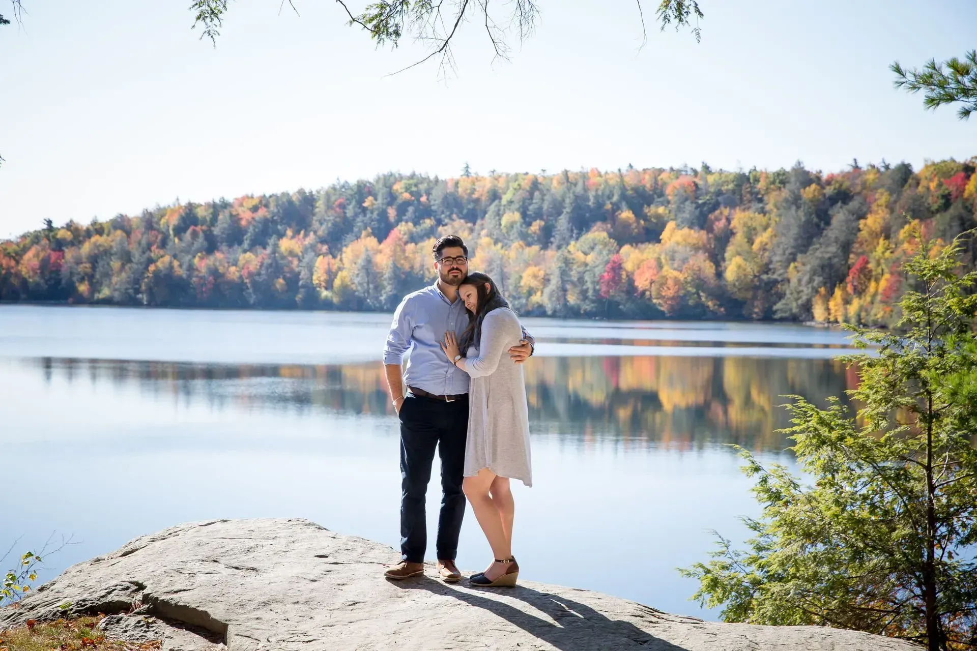 Couple hugging by a lake, autumn foliage in background. Sunny day, soft colors.