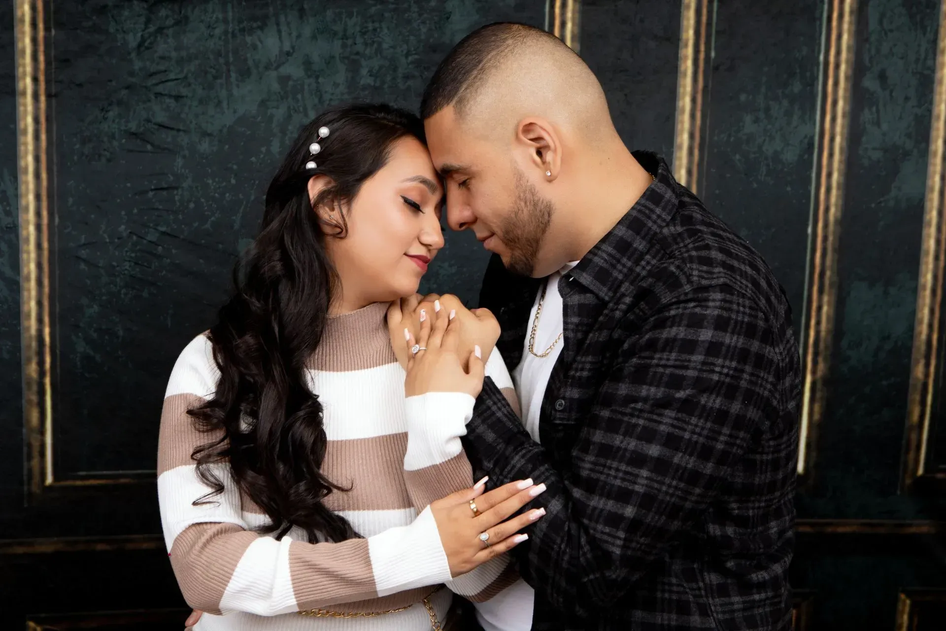 Couple embraces, holding hands with foreheads touching. Dark plaid shirt, striped sweater, against dark background.