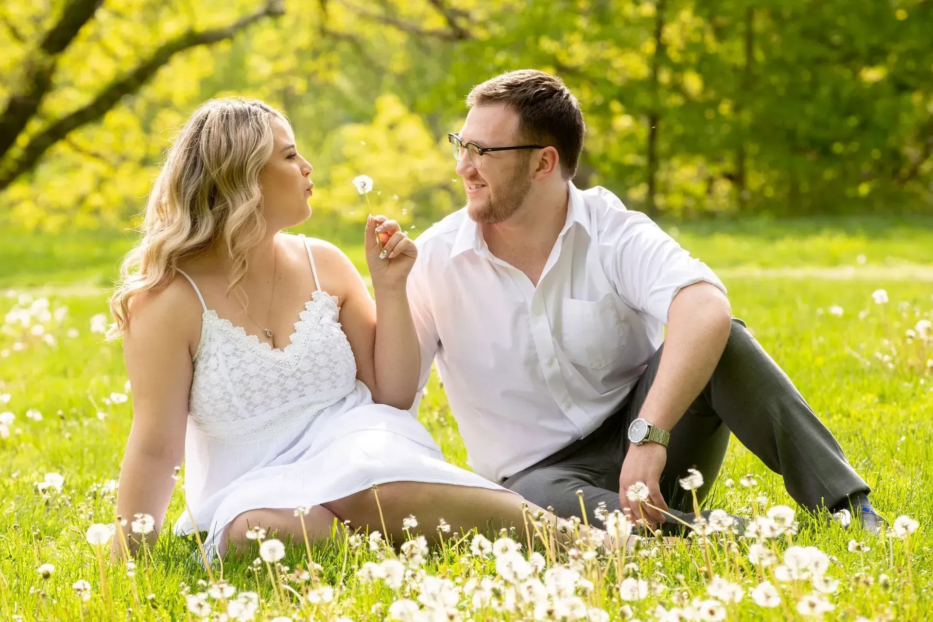 Couple seated in a field of flowers, woman blowing dandelion towards man who smiles.