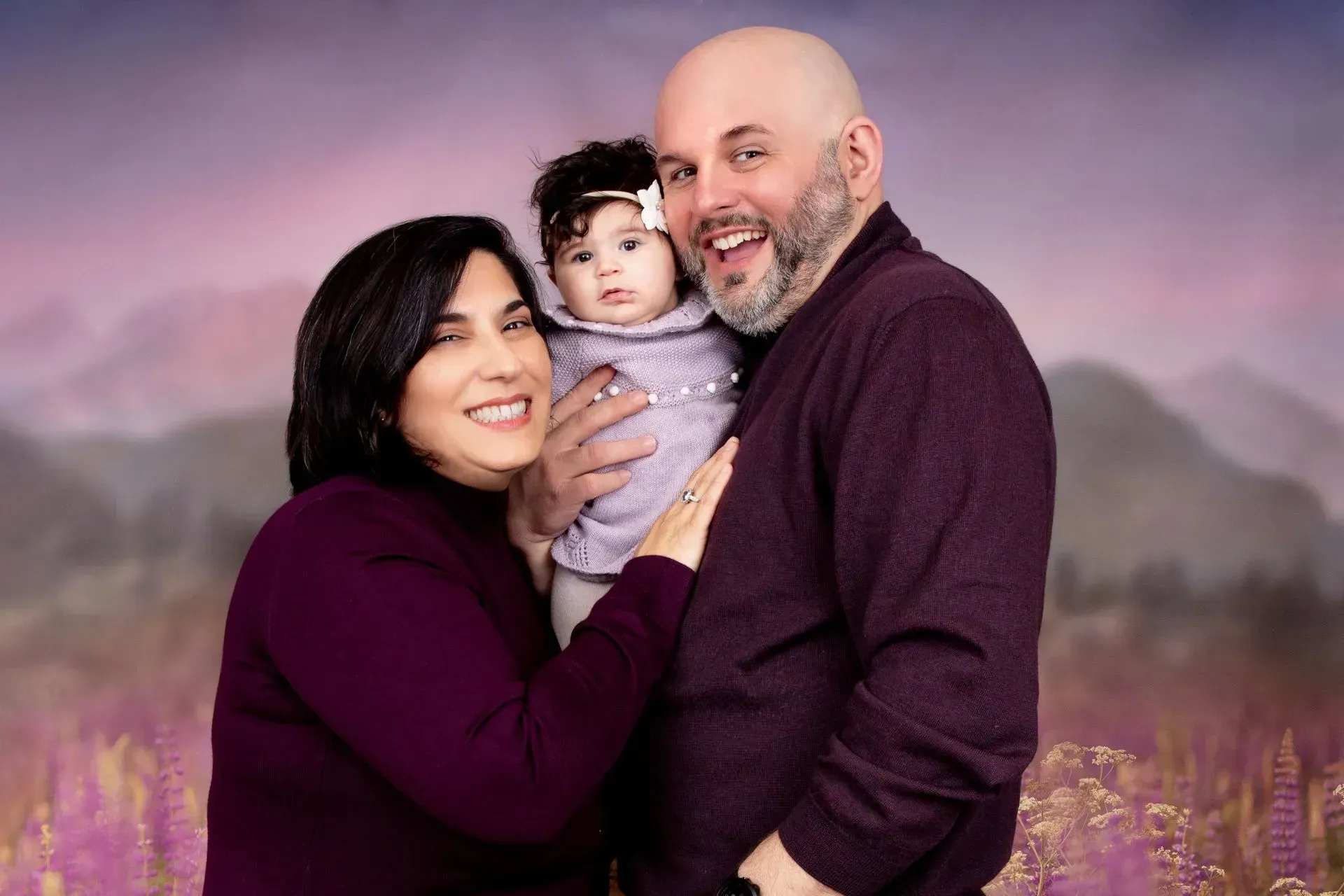 Family of three in matching purple sweaters, posing in front of a painted landscape with purple flowers.