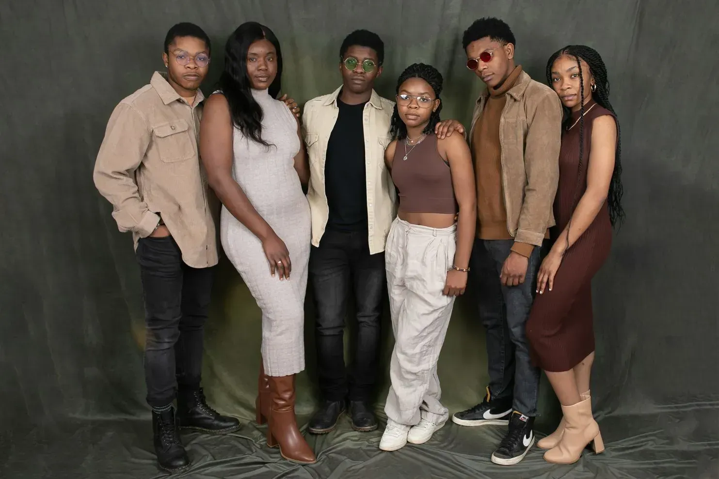 Six people pose together, mostly standing, against a dark backdrop. They wear various neutral-toned outfits, some with jackets and sunglasses.