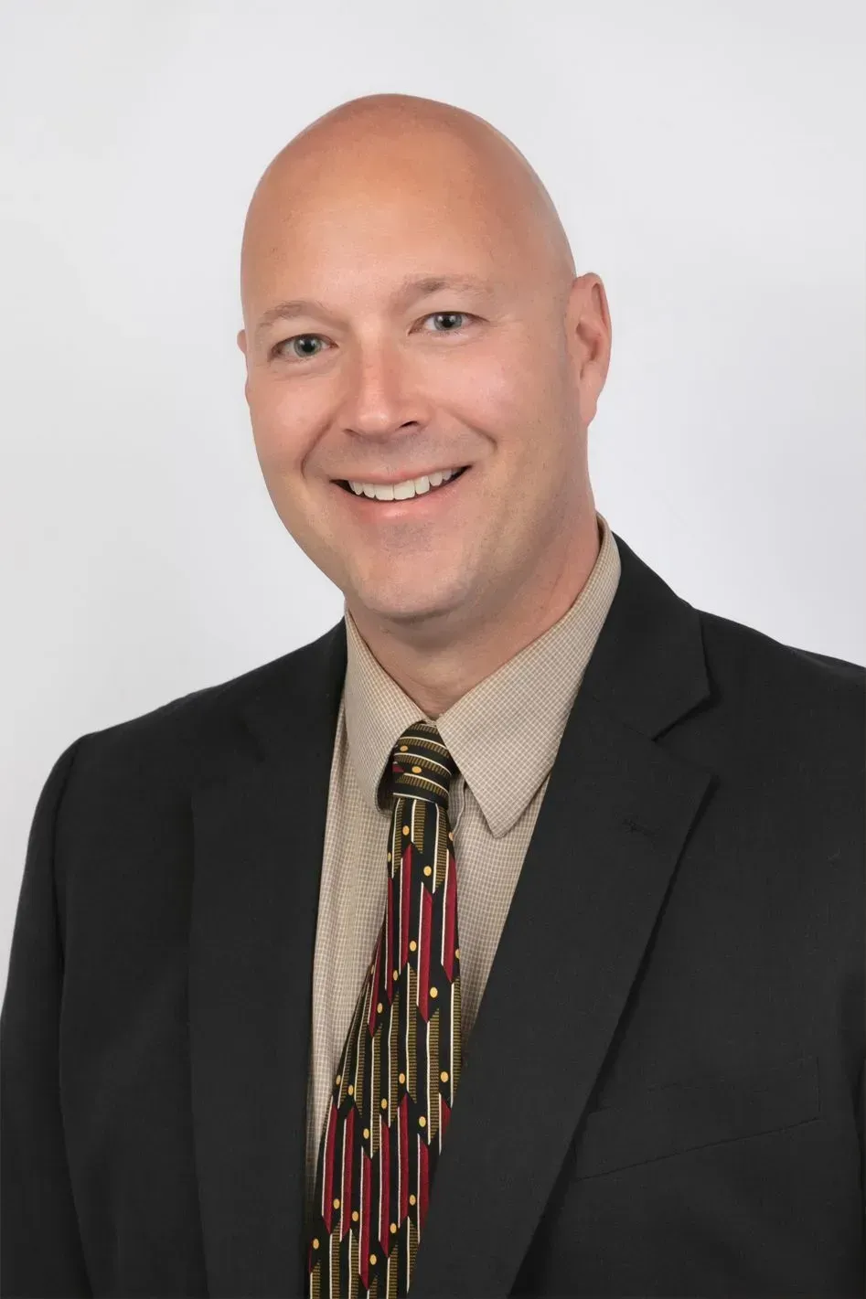 Bald man smiles wearing a suit and patterned tie against a white background.