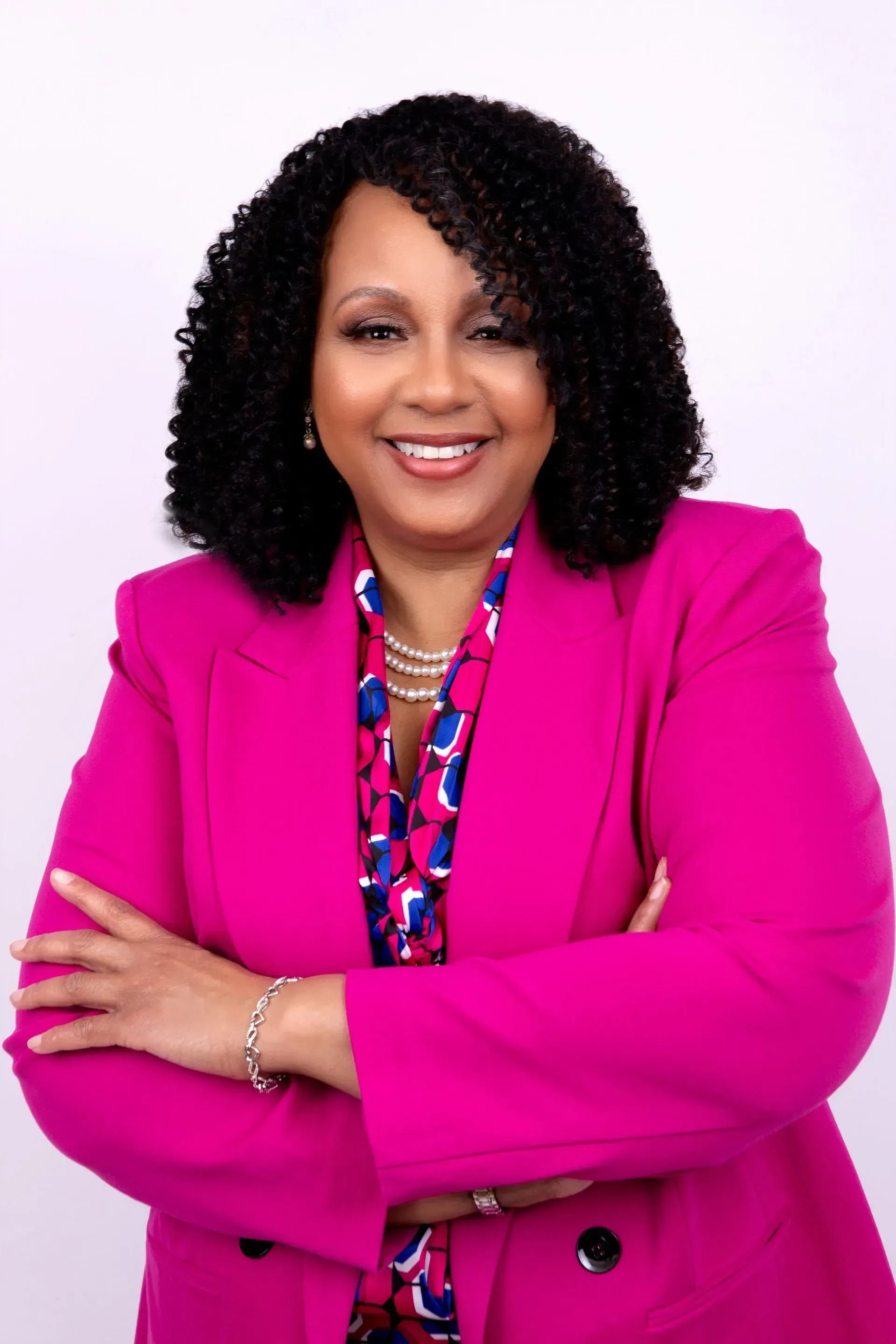 Woman with dark curly hair, wearing a pink blazer, smiles with arms crossed. White background.