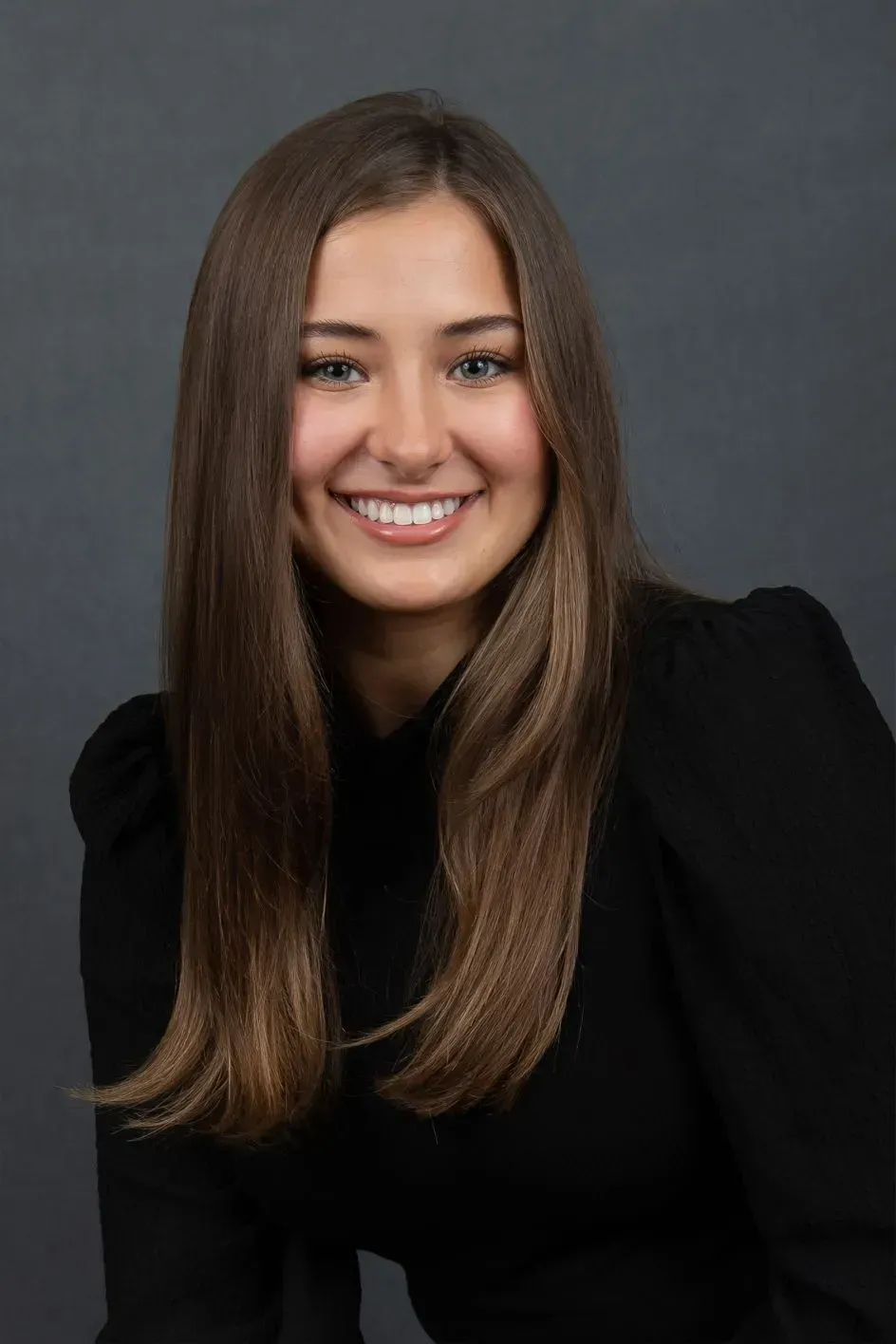 Woman smiling at the camera, wearing a black top, with long brown hair, against a dark gray background.