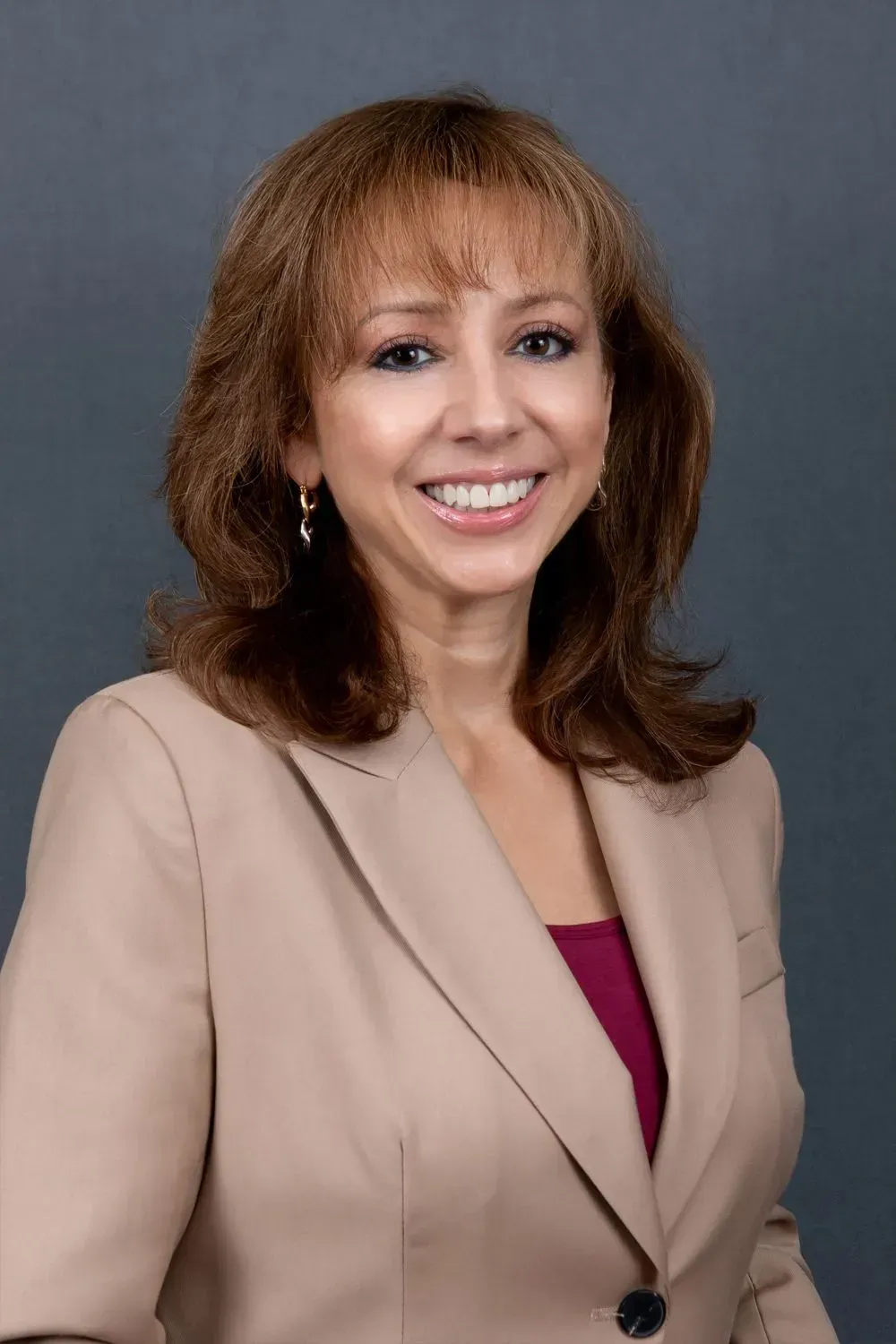 Woman smiling, wearing a tan blazer over a burgundy top, standing against a gray backdrop.