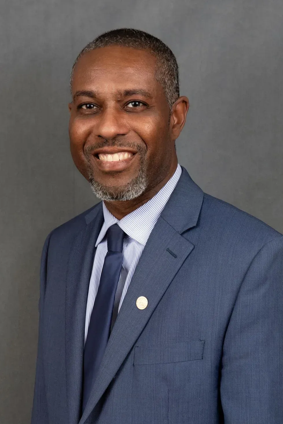 Man in blue suit, smiling, wearing tie and small gold emblem on lapel. Gray background.