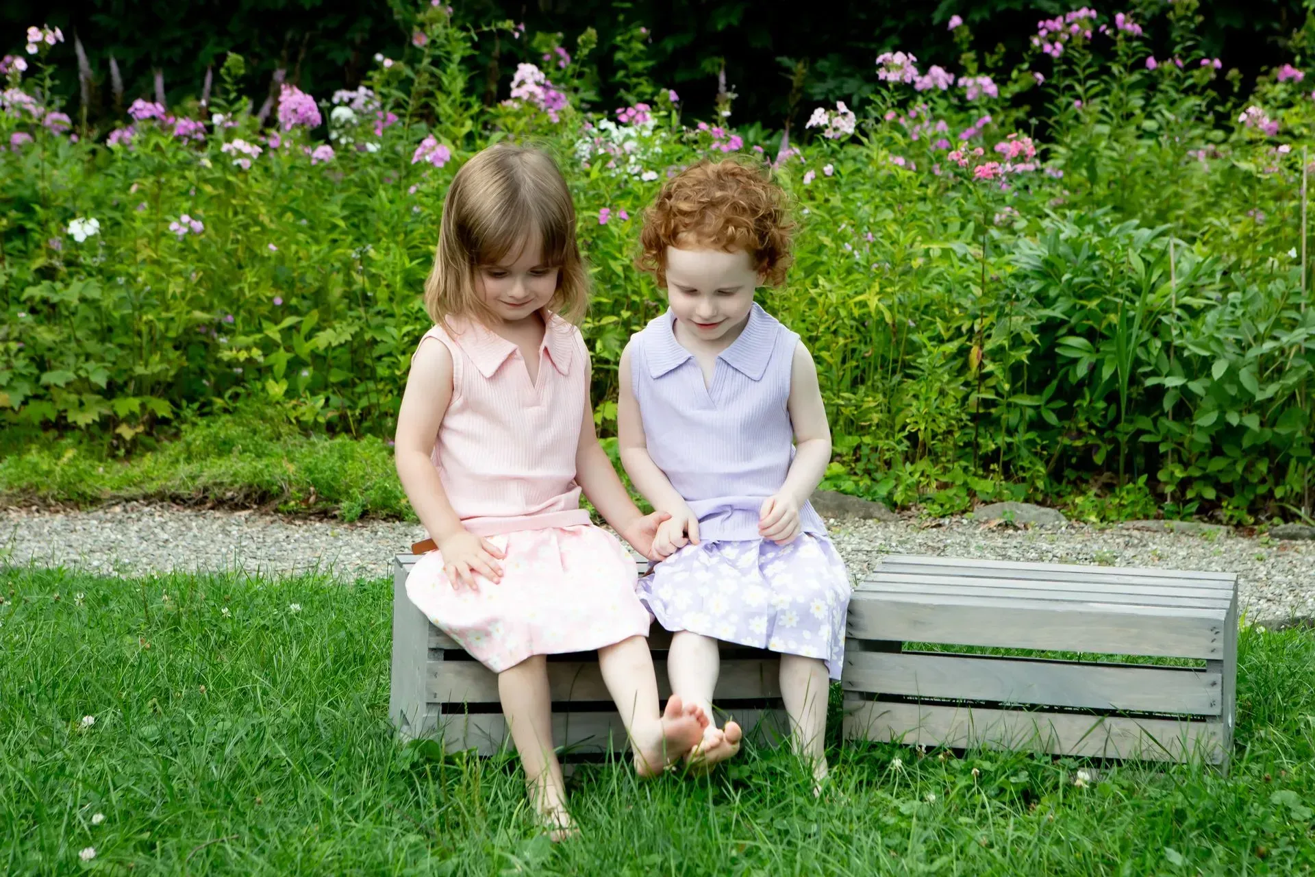 Two young girls in dresses sit holding hands on a wooden crate in a garden.