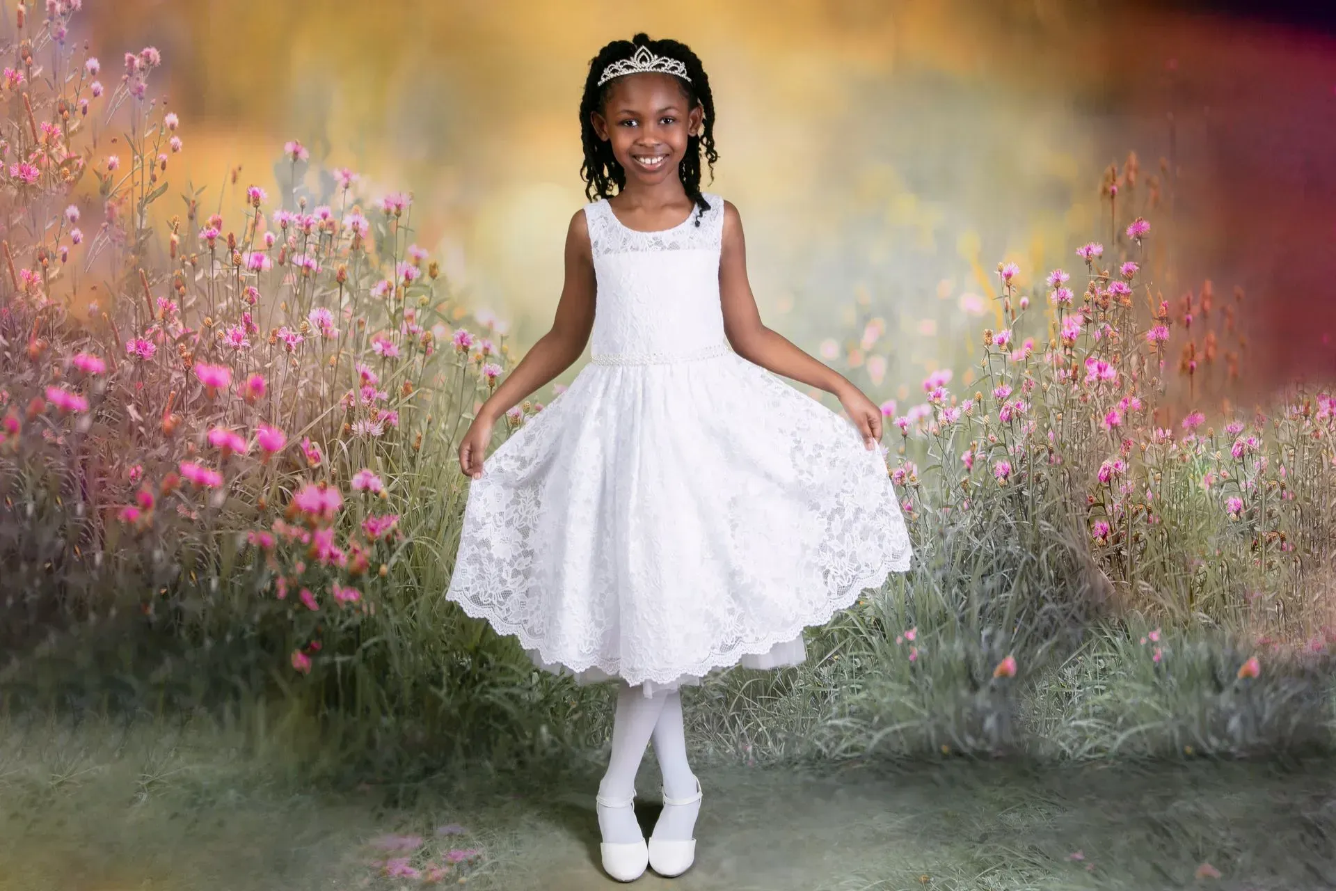 Girl in white lace dress and headband smiles, holding dress. Standing in a field of flowers.