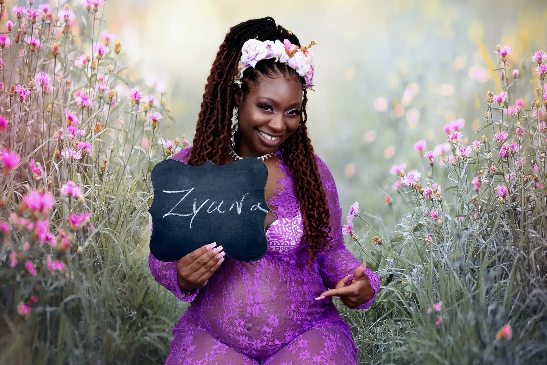 Pregnant person in a lavender lace outfit holds a sign that says Zyda in a field of flowers and points to her belly.