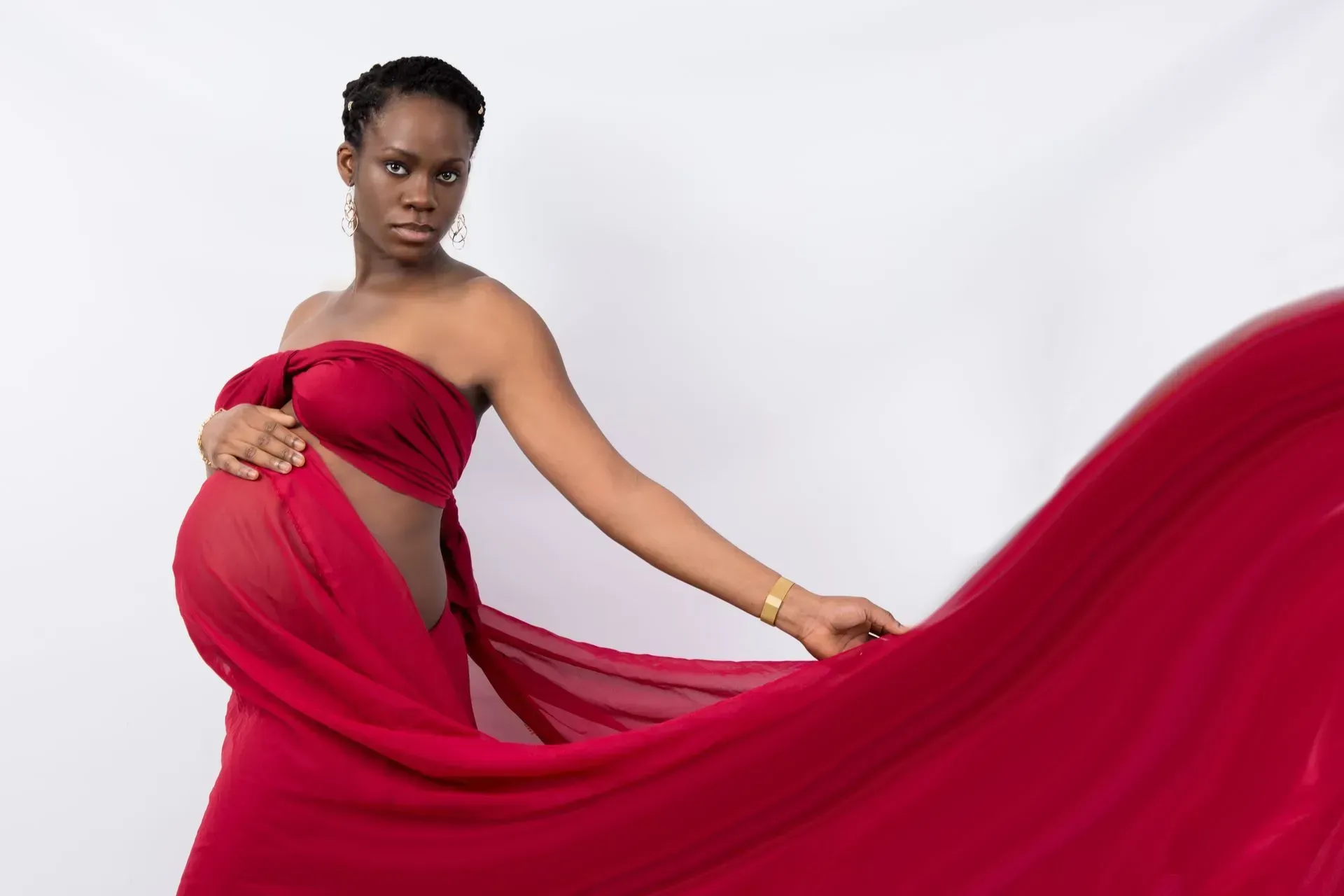 Pregnant person in red draped fabric, hands on belly, against white backdrop.