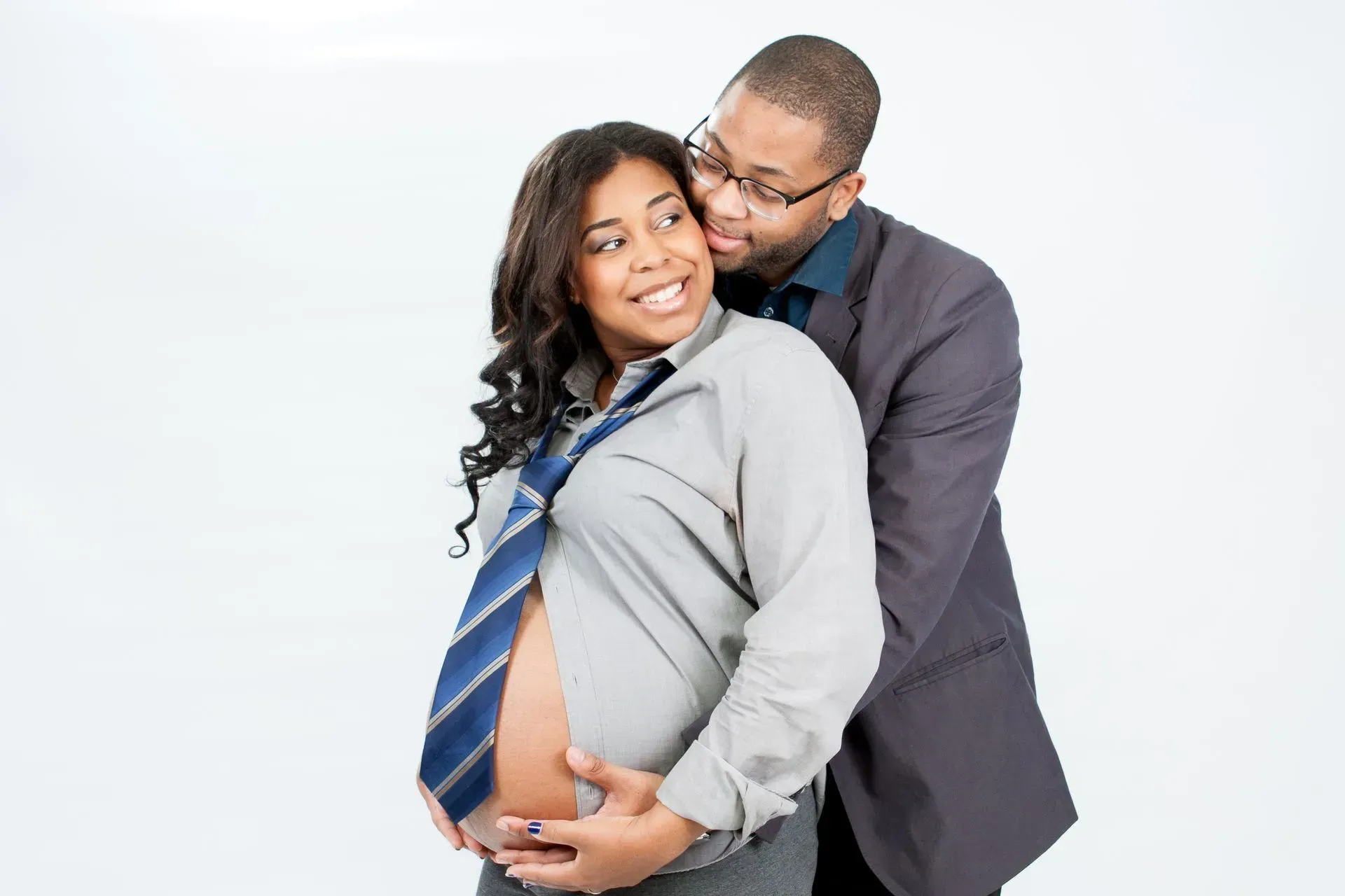 Man hugging and kissing pregnant woman, both smiling, studio shot. Woman wearing shirt and tie, man in suit jacket.