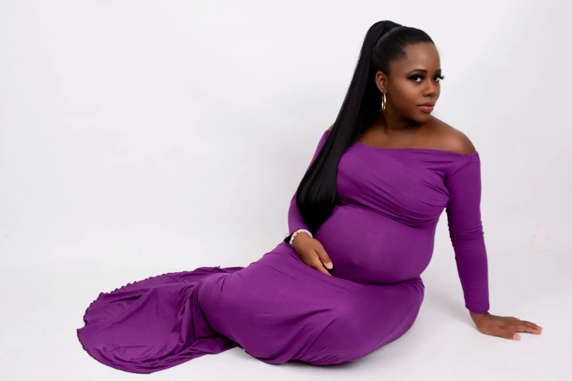 Pregnant person in a purple off-the-shoulder dress, sitting on the floor, hand on belly, looking at the camera.