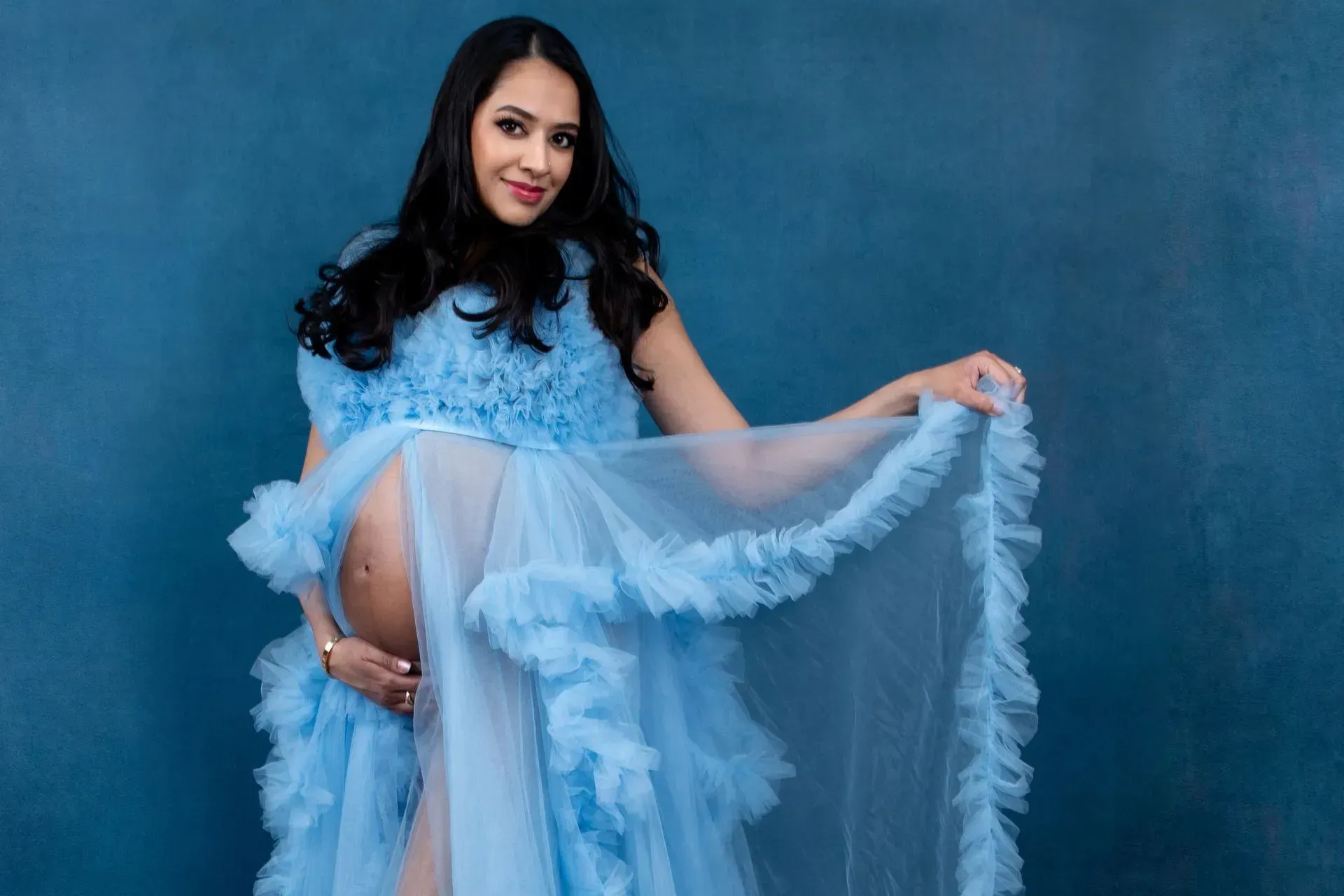 Pregnant person in light blue ruffled dress, holding belly, against a blue backdrop.