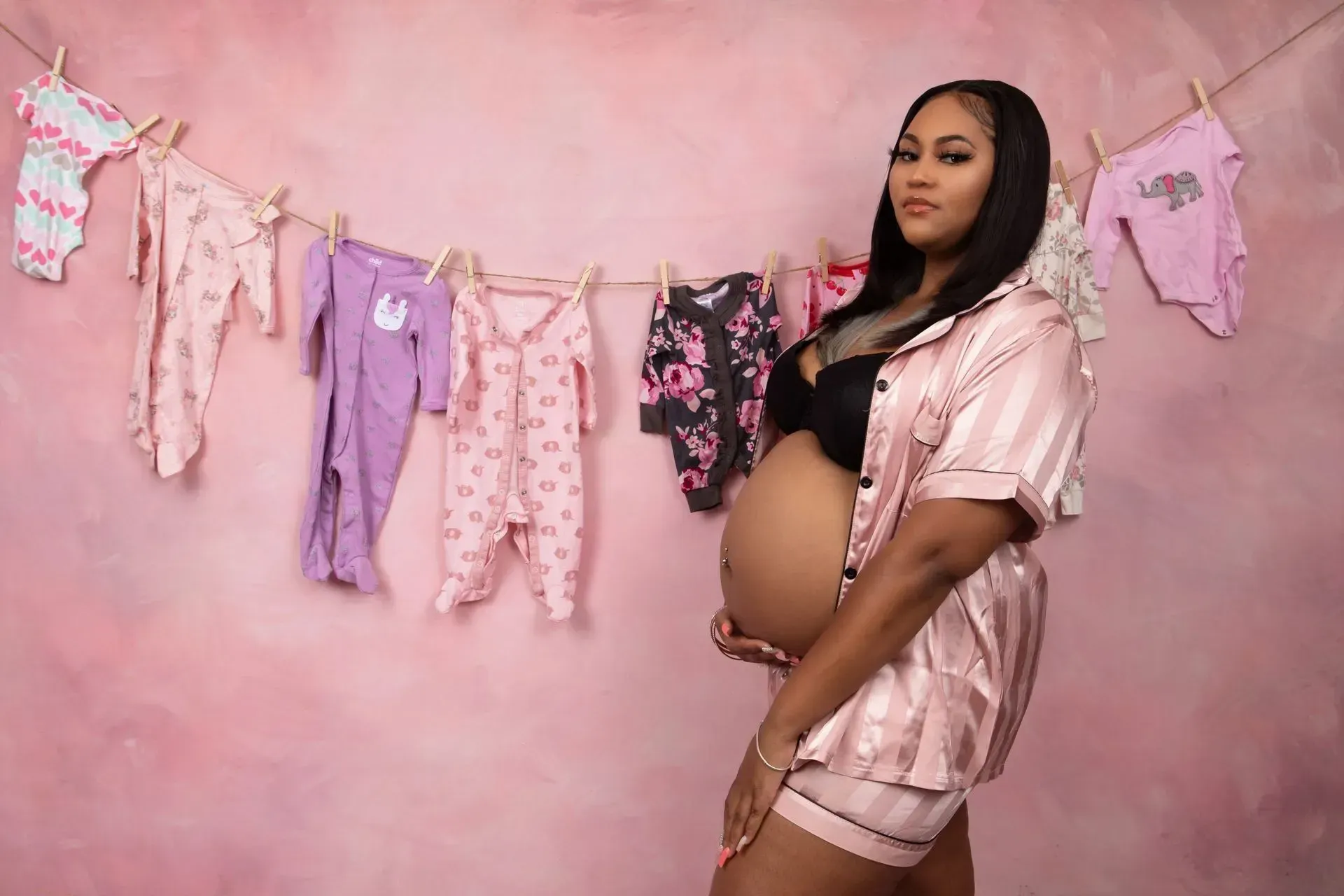 Pregnant person in pink pajamas, holding belly, posing in front of baby clothes hung on a line. Pink background.