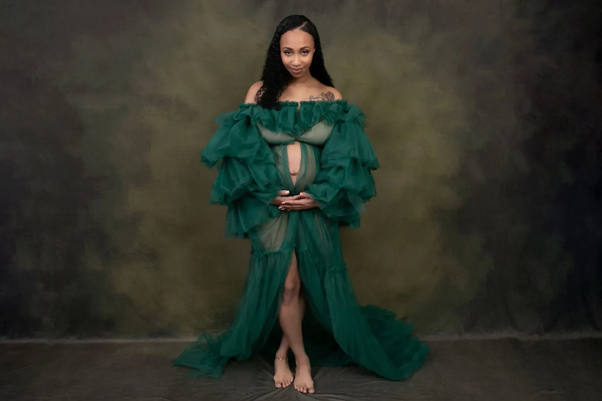 Pregnant person in a green gown, hands on belly, standing in front of an abstract, dark background.