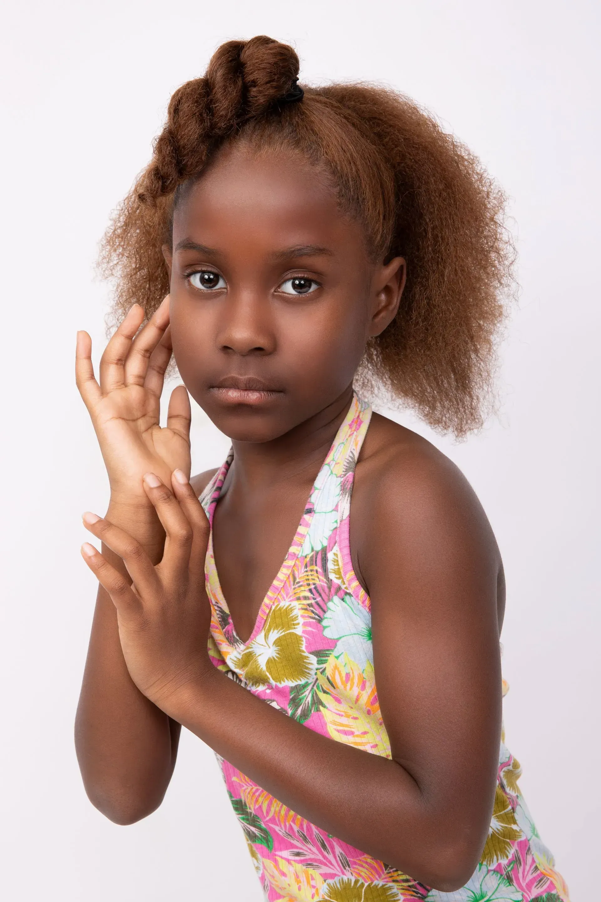 Young person with brown skin and styled hair, wearing a floral halter top, posing with hands near face.