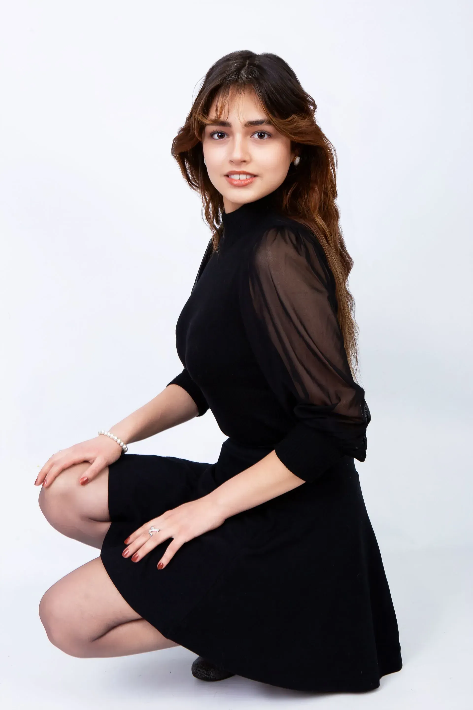 Woman in a black dress, crouched, looking at the camera, against a white backdrop.