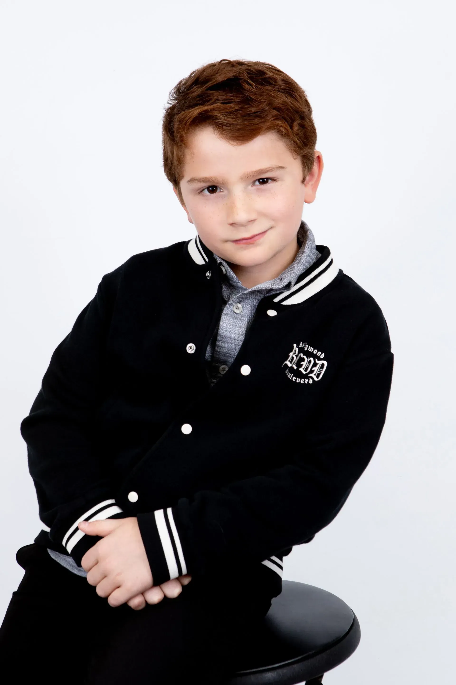 Boy with red hair in a black letterman jacket, seated with a slight smile.