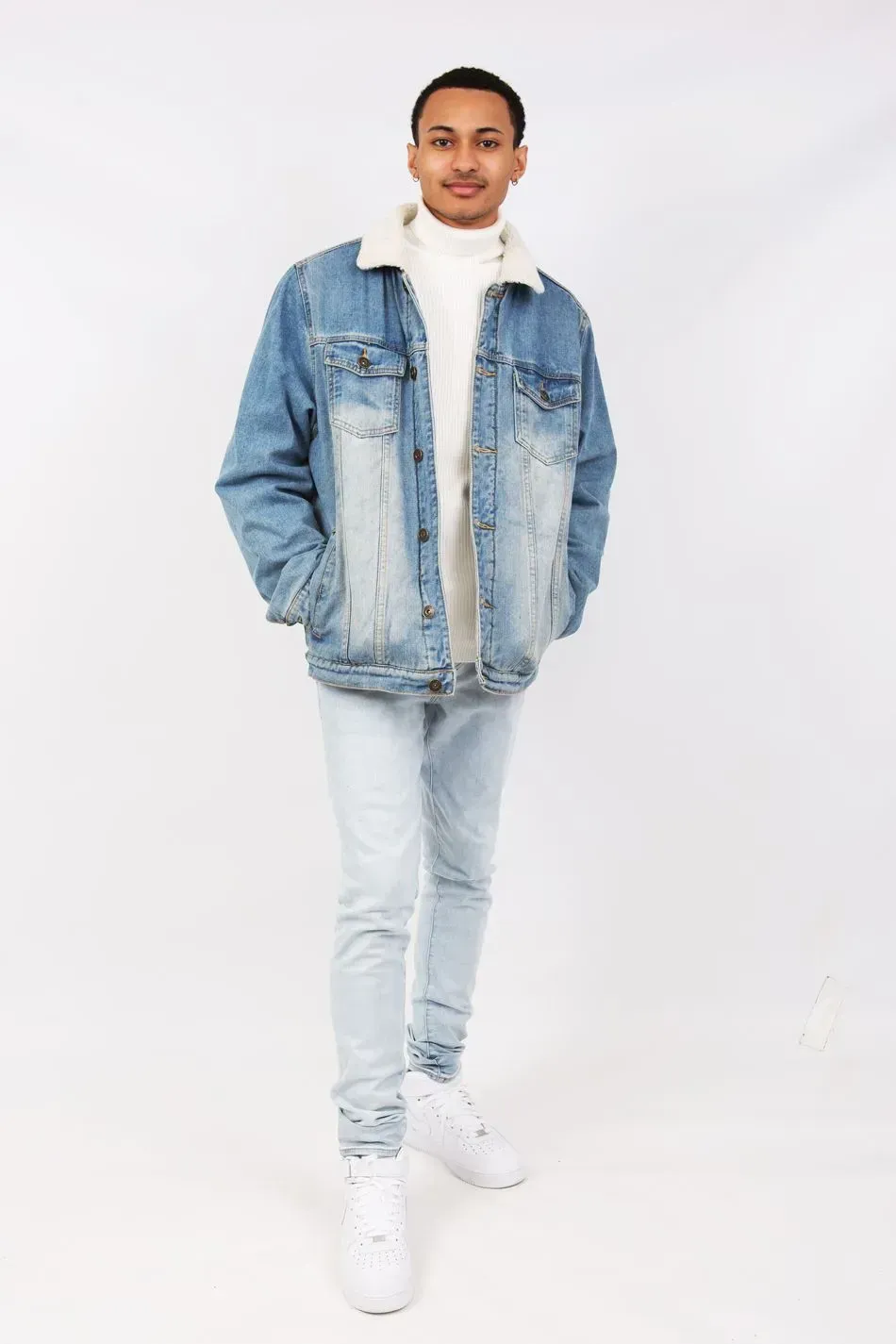 Man in denim jacket with white turtleneck and light jeans, hands in pockets.