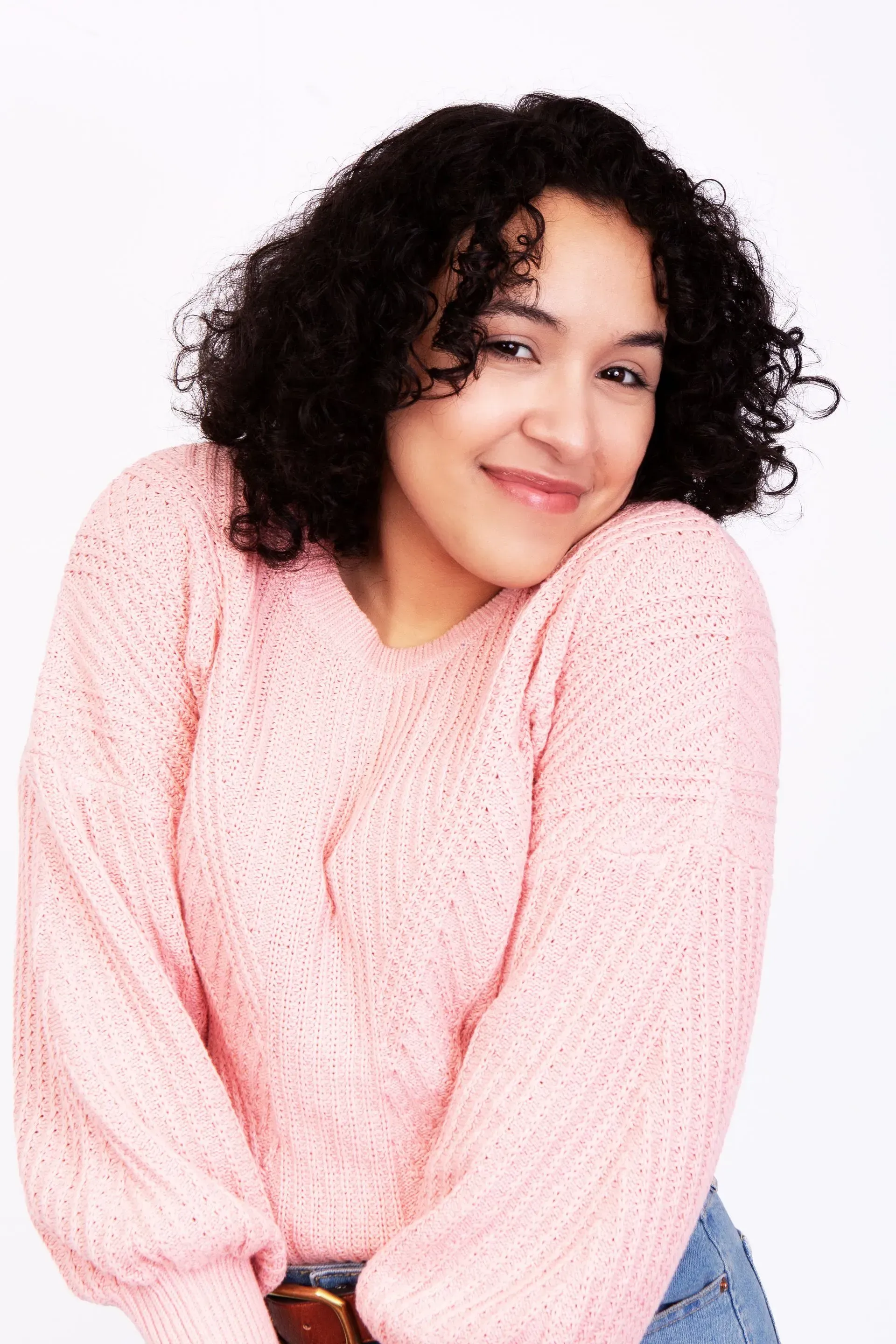 Woman in pink sweater smiling, curly hair, leaning forward. White background.
